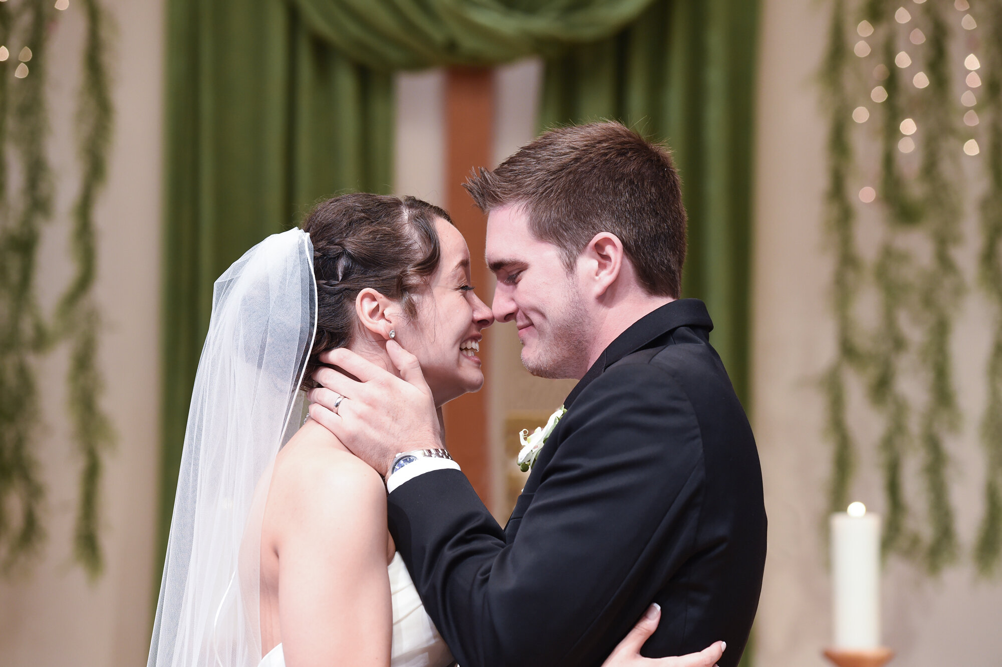 Bride and groom embracing and smiling during wedding ceremony, with green drapes in the background.