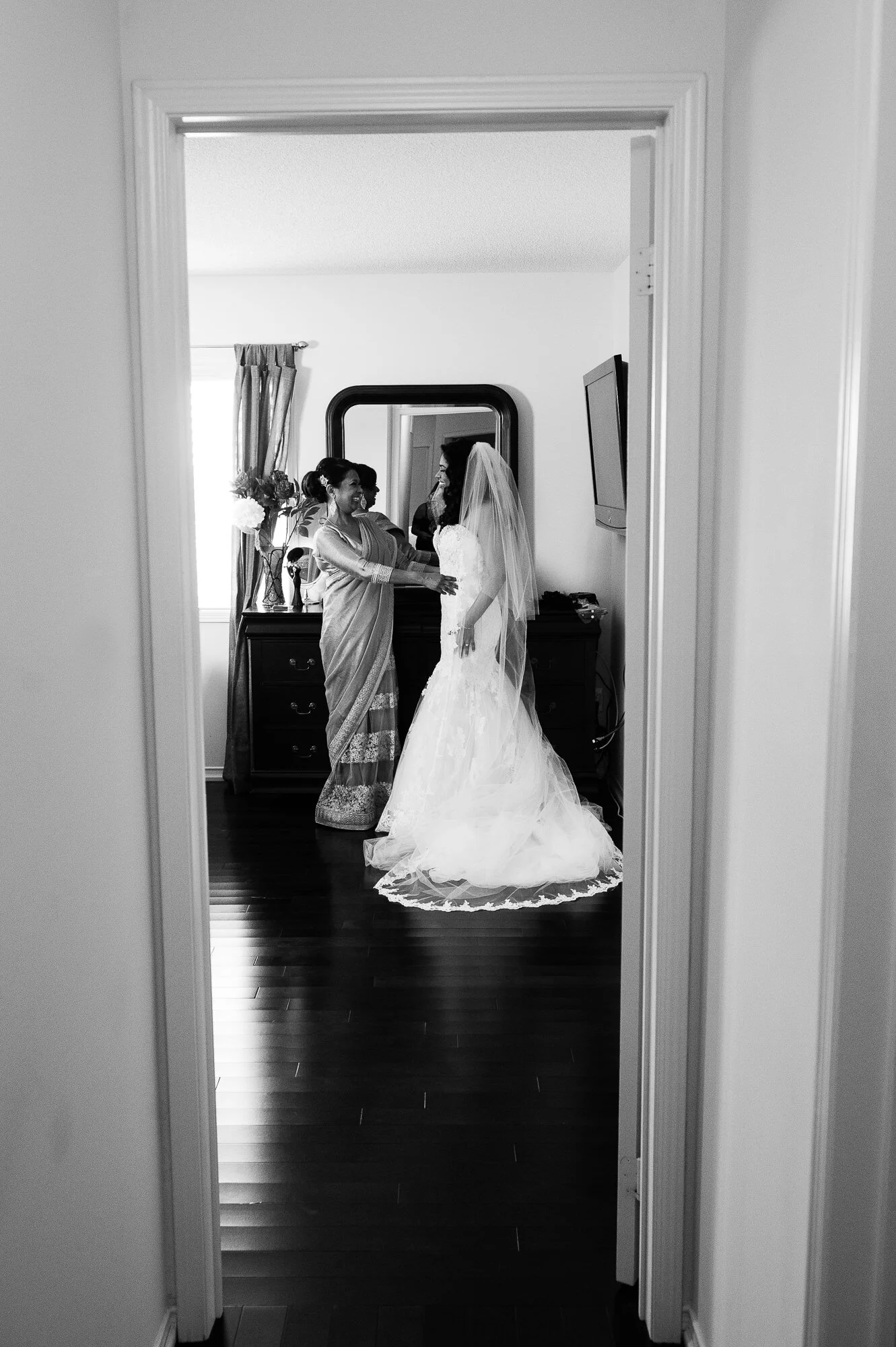 Black and white photo of a bride in a gown and veil in a bedroom, being assisted by another woman in a sari. They are standing near a dresser with a mirror.