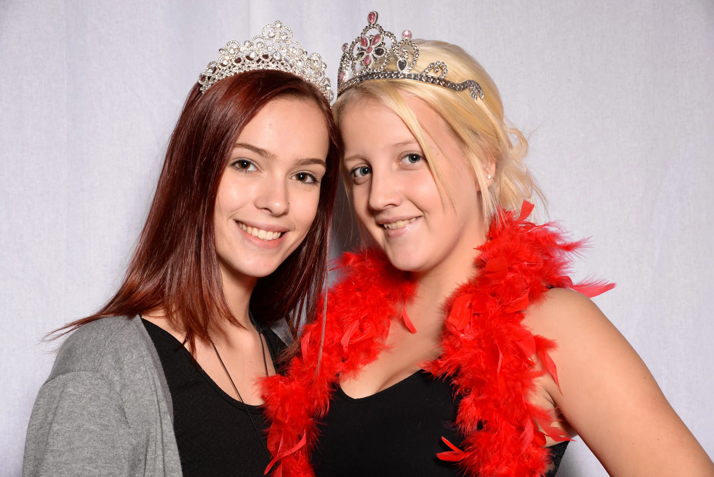 Two young women smiling, wearing tiaras; one with a red feather boa.