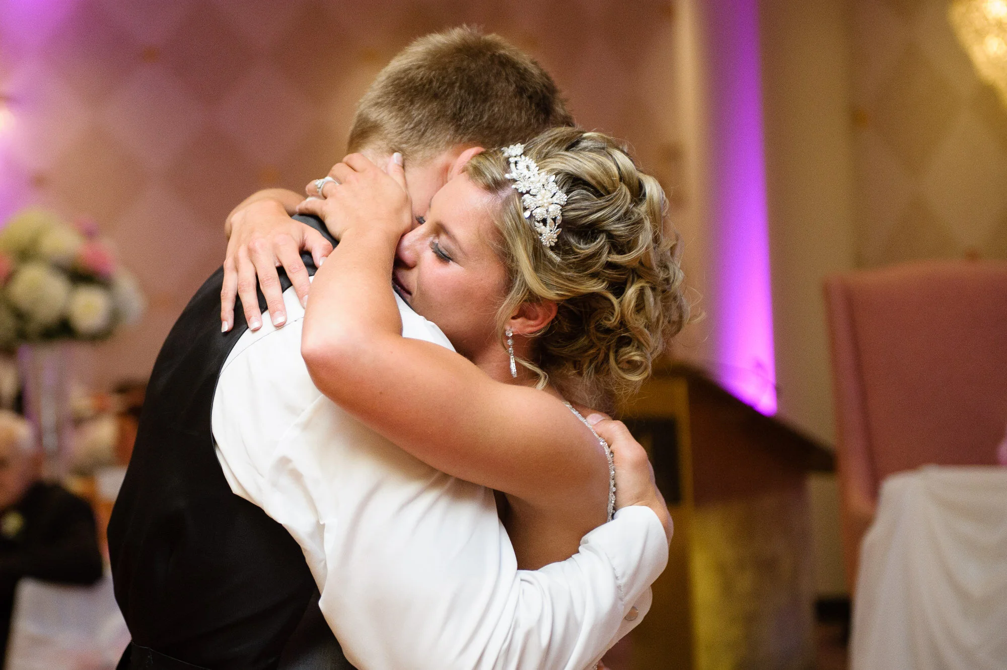 A bride and groom hugging during a wedding reception, with the bride wearing a jeweled hair accessory and elegant earrings.