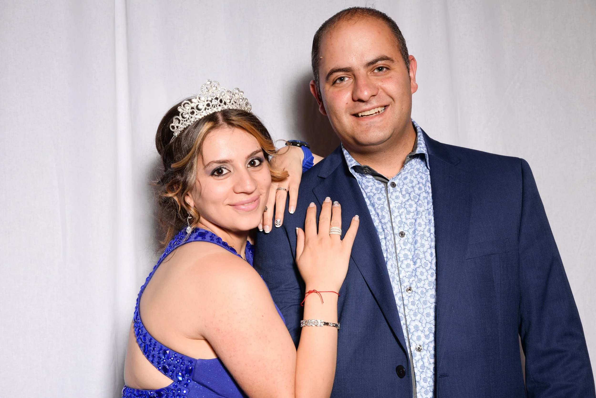 Man and woman posing together, woman wearing a tiara and blue dress, man in a blue suit, against a white curtain backdrop.