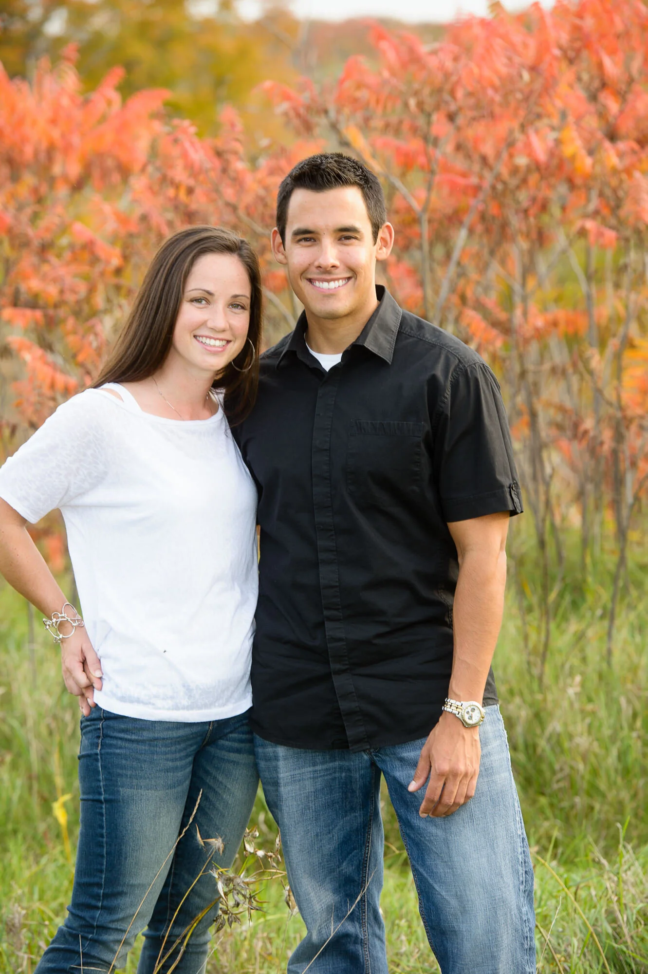 A smiling couple standing together in a grassy field with red-orange foliage in the background. The woman is wearing a white shirt and jeans, while the man is wearing a black shirt and jeans.