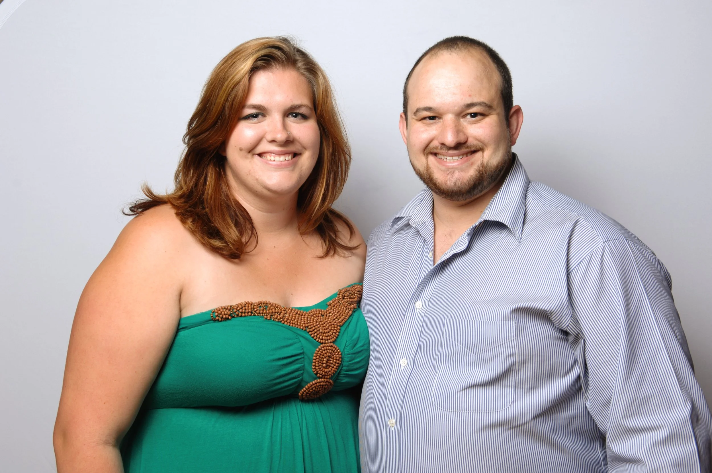 A man and woman smiling, standing side by side. The woman is wearing a green strapless dress with a decorative pattern, and the man is in a blue striped button-down shirt. The background is light gray.