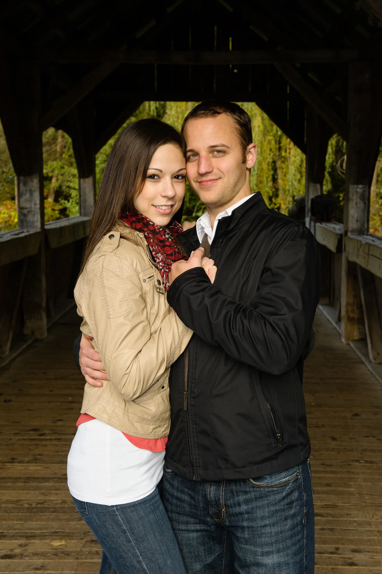 A couple posing together in a covered wooden bridge with the man wearing a black jacket and the woman in a beige jacket and red scarf.