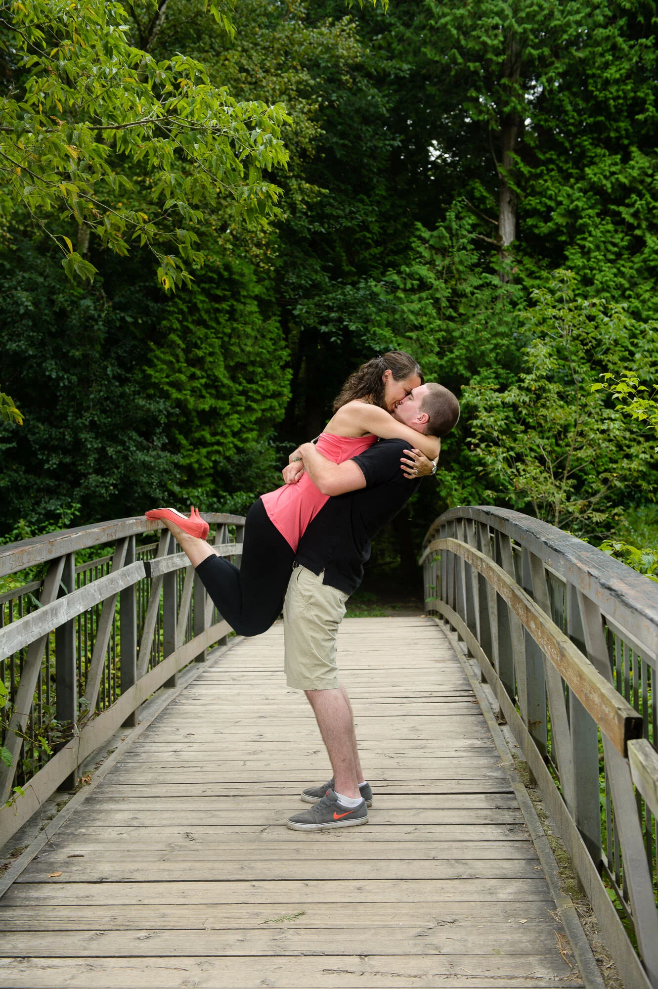 A couple sharing a romantic kiss on a wooden bridge surrounded by lush greenery.