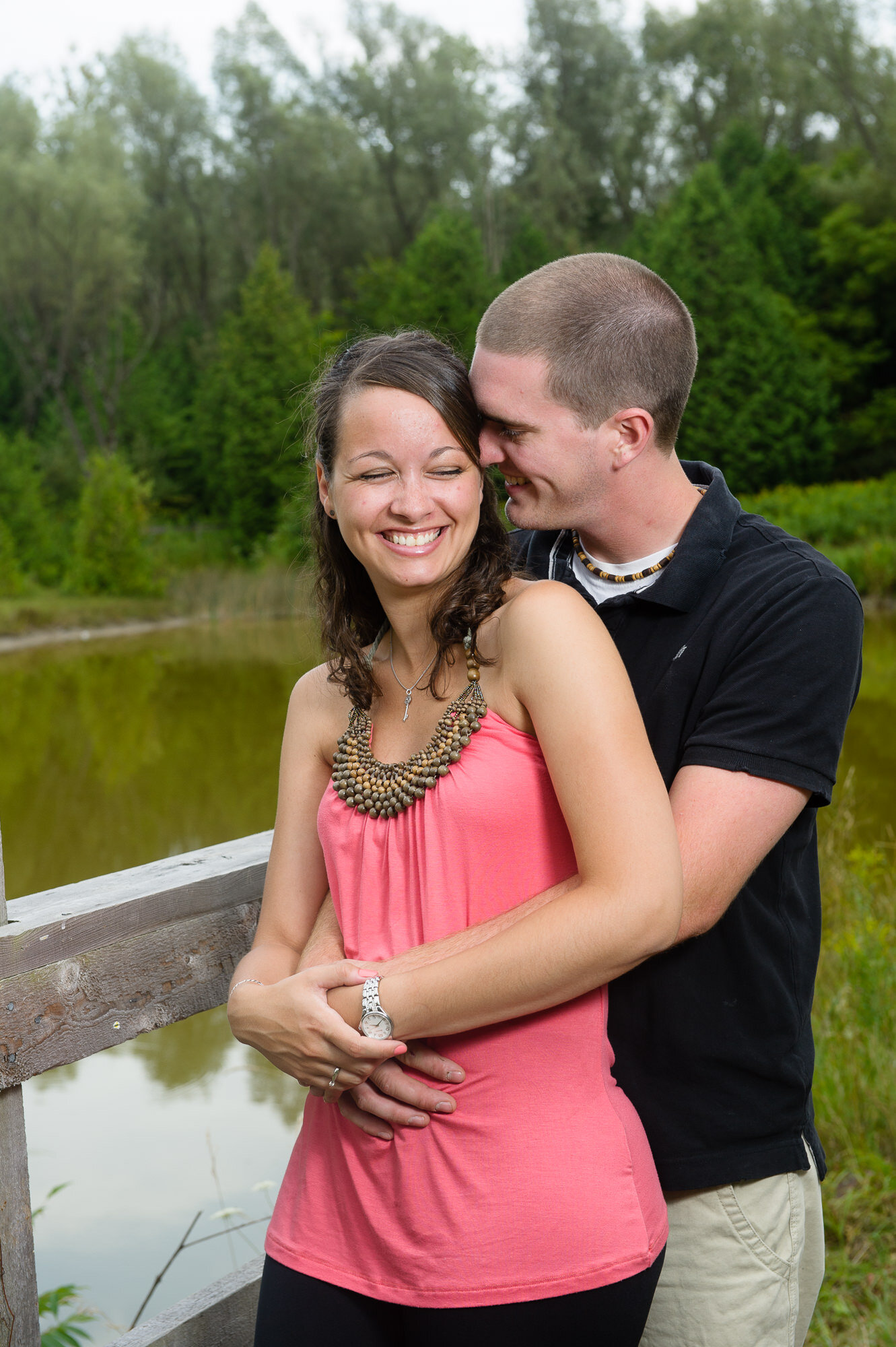 A couple embracing on a wooden bridge near a pond with trees in the background. The woman is wearing a pink top and the man a black shirt.