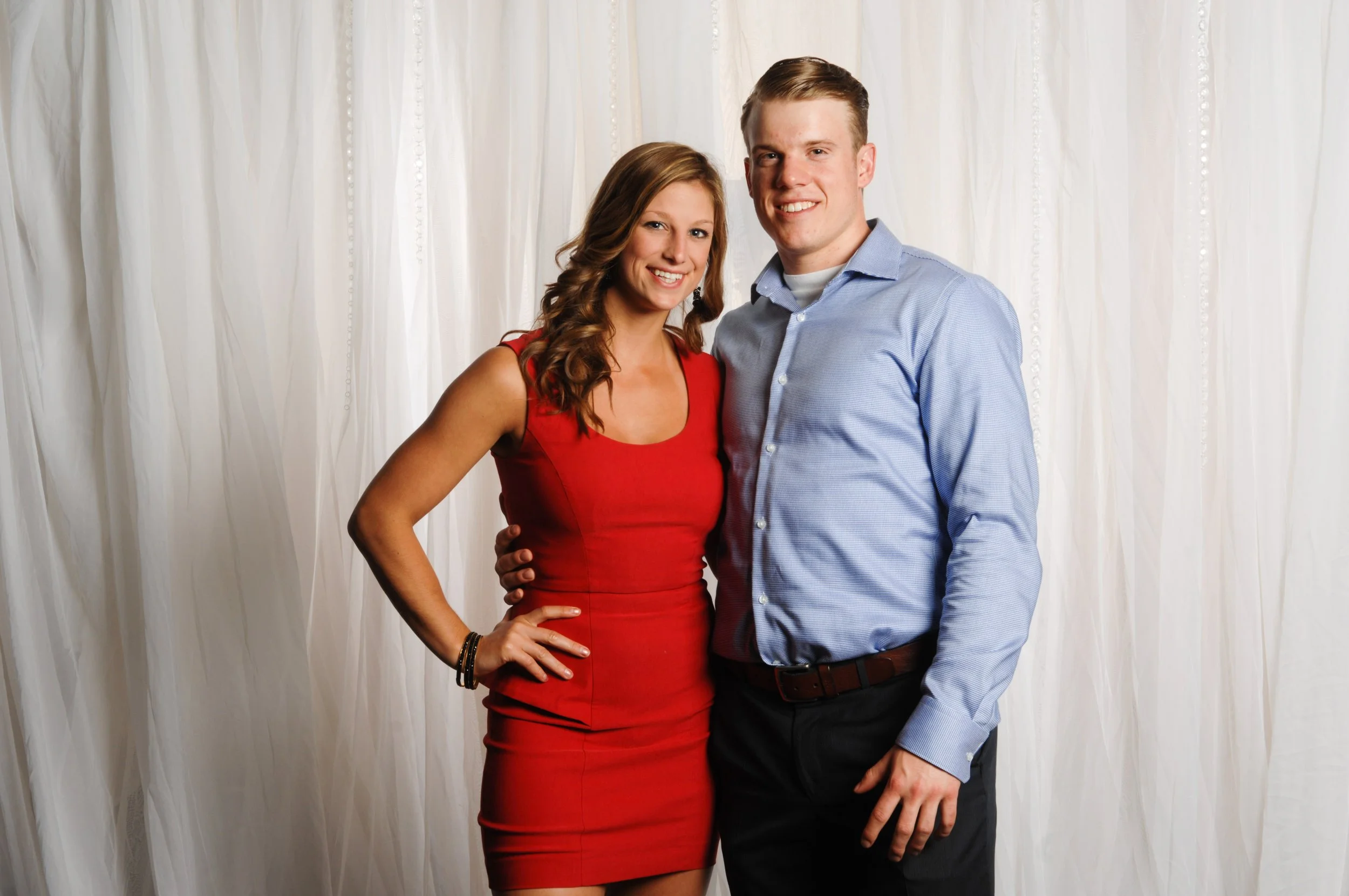 A man in a blue shirt and a woman in a red dress posing in front of a white curtain background.