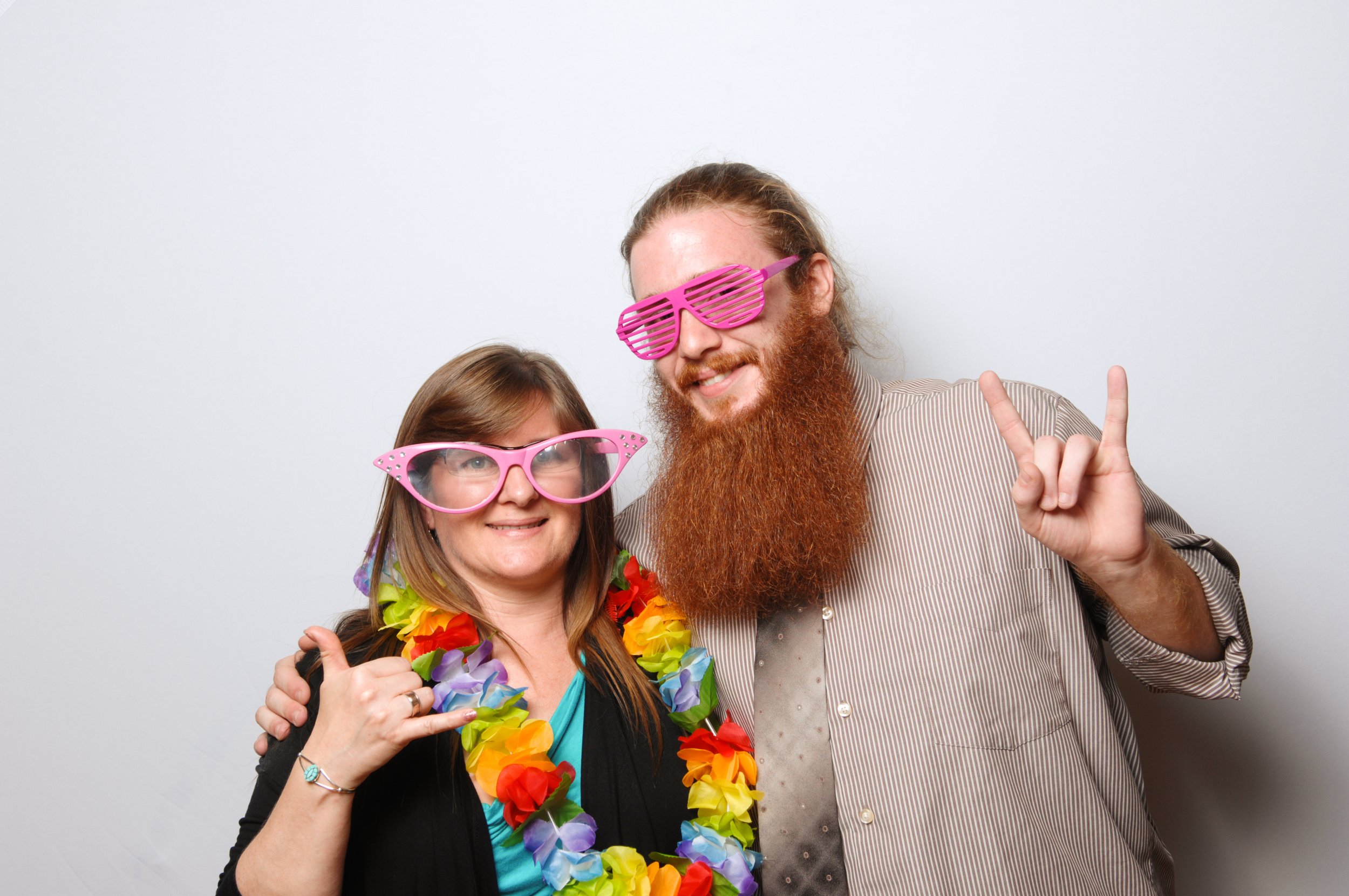Two people posing playfully with oversized pink glasses and a colorful lei, making fun hand gestures.
