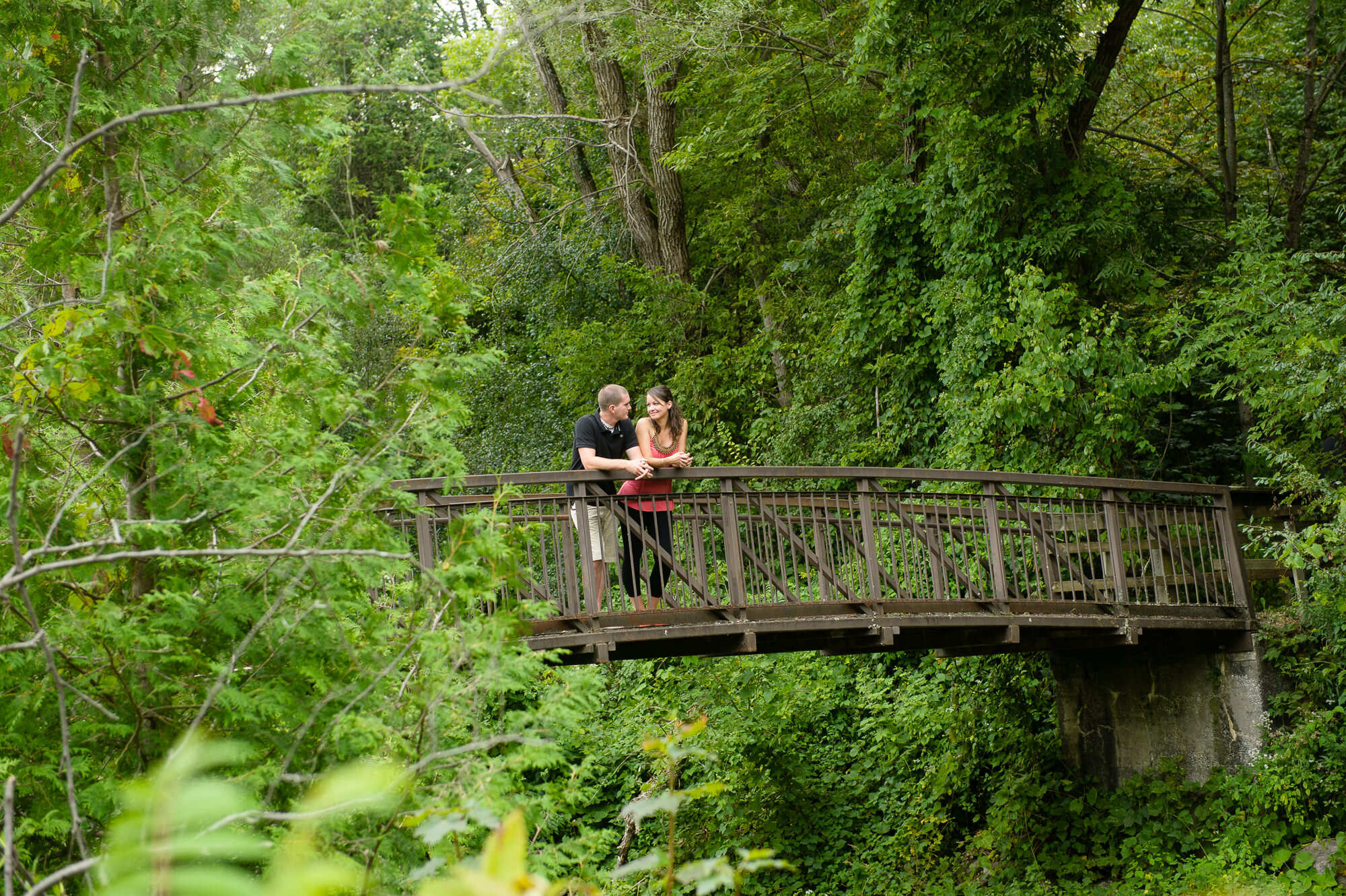 A couple standing on a wooden bridge surrounded by lush green trees.