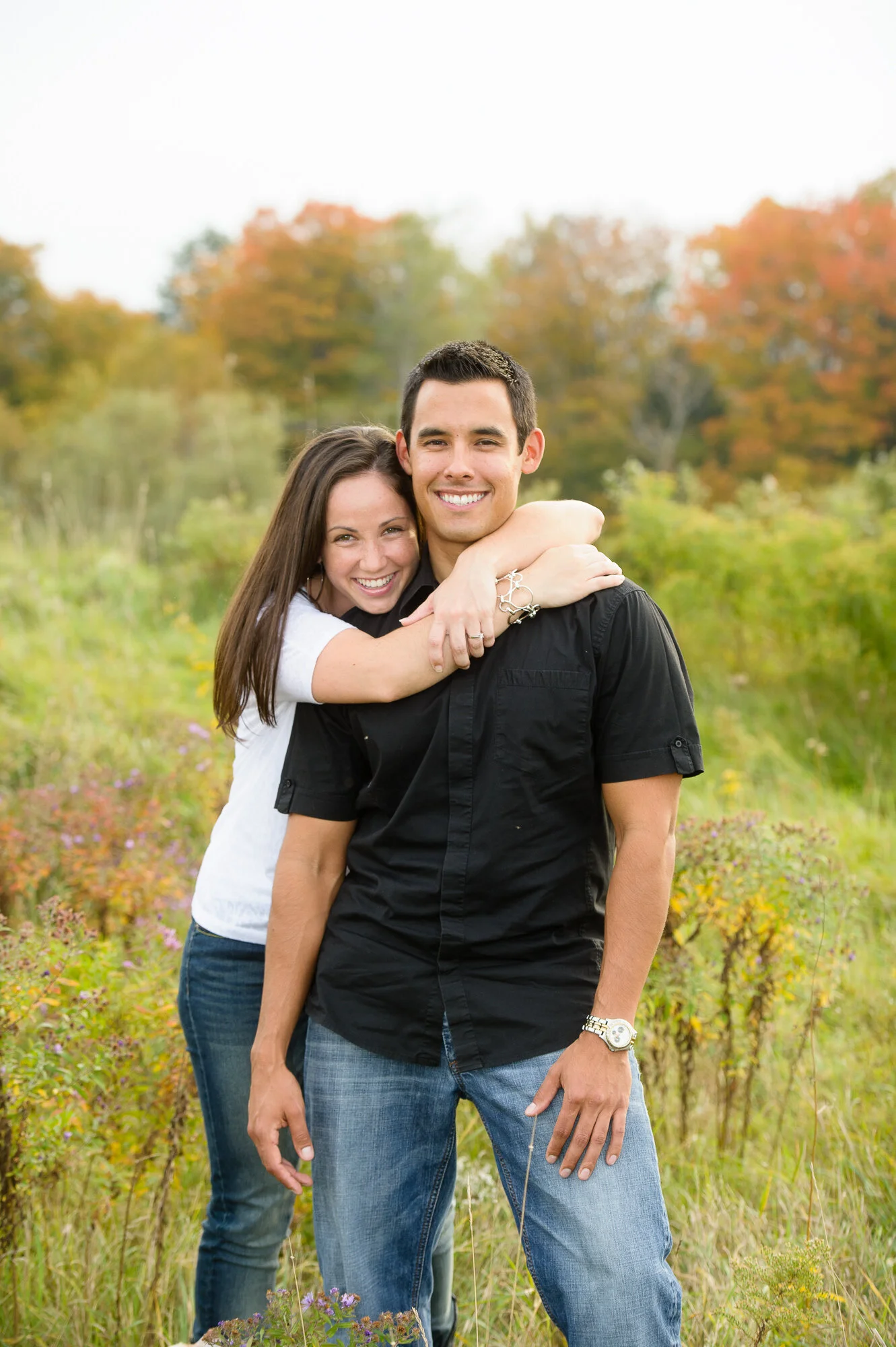 Smiling couple embracing in a grassy field with autumn trees in the background.