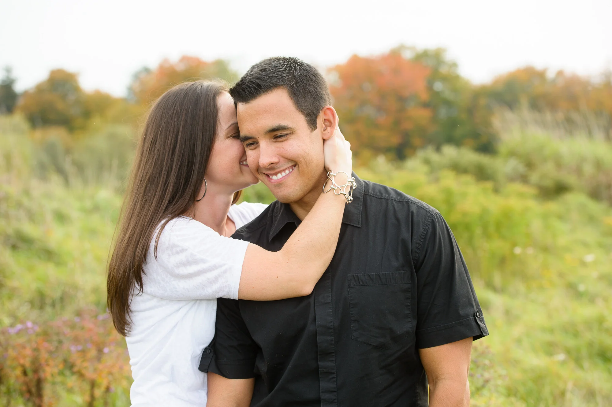 A woman whispering to a smiling man in a field with autumn foliage in the background.