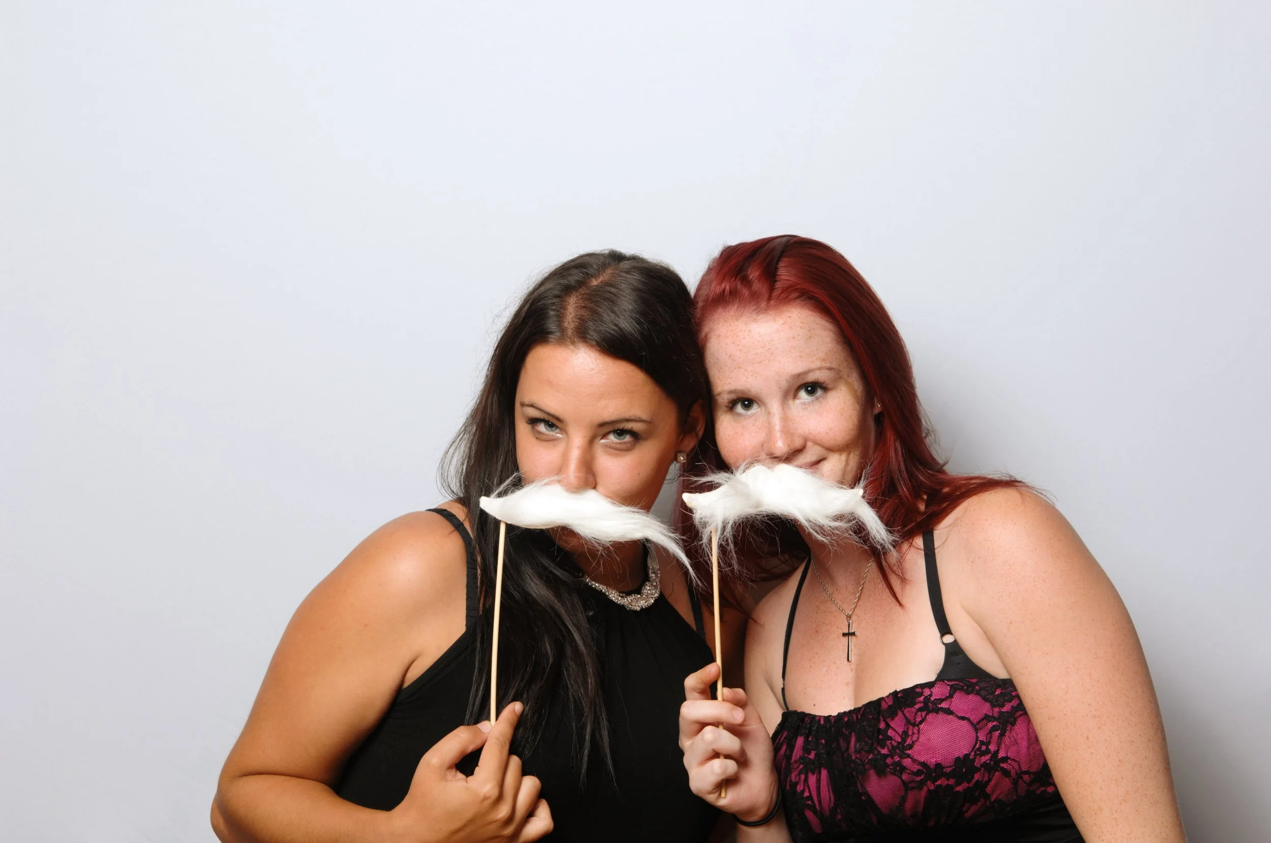 Two women holding white fake mustaches on sticks, smiling playfully against a plain background.