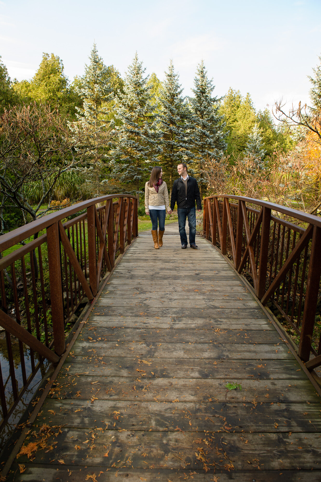 A couple holding hands while walking on a wooden bridge surrounded by trees in a park or natural setting.