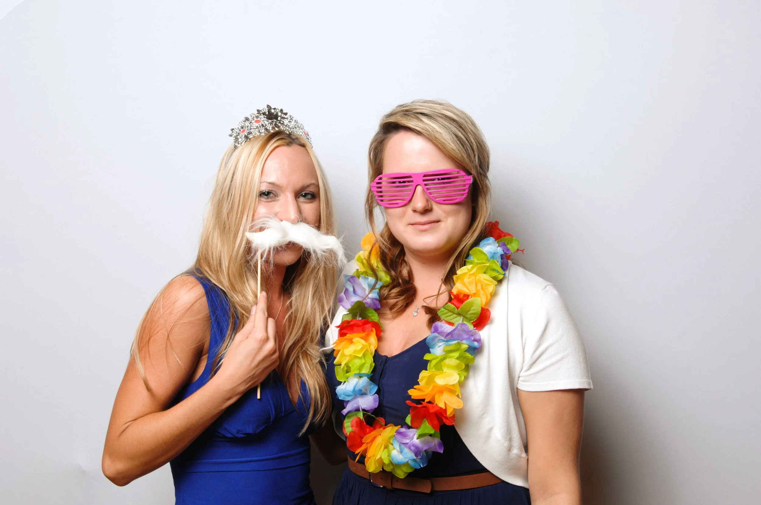 Two women in a playful pose, one wearing a tiara and fake white mustache, the other with pink shutter shades and a colorful lei; both smiling against a plain background.