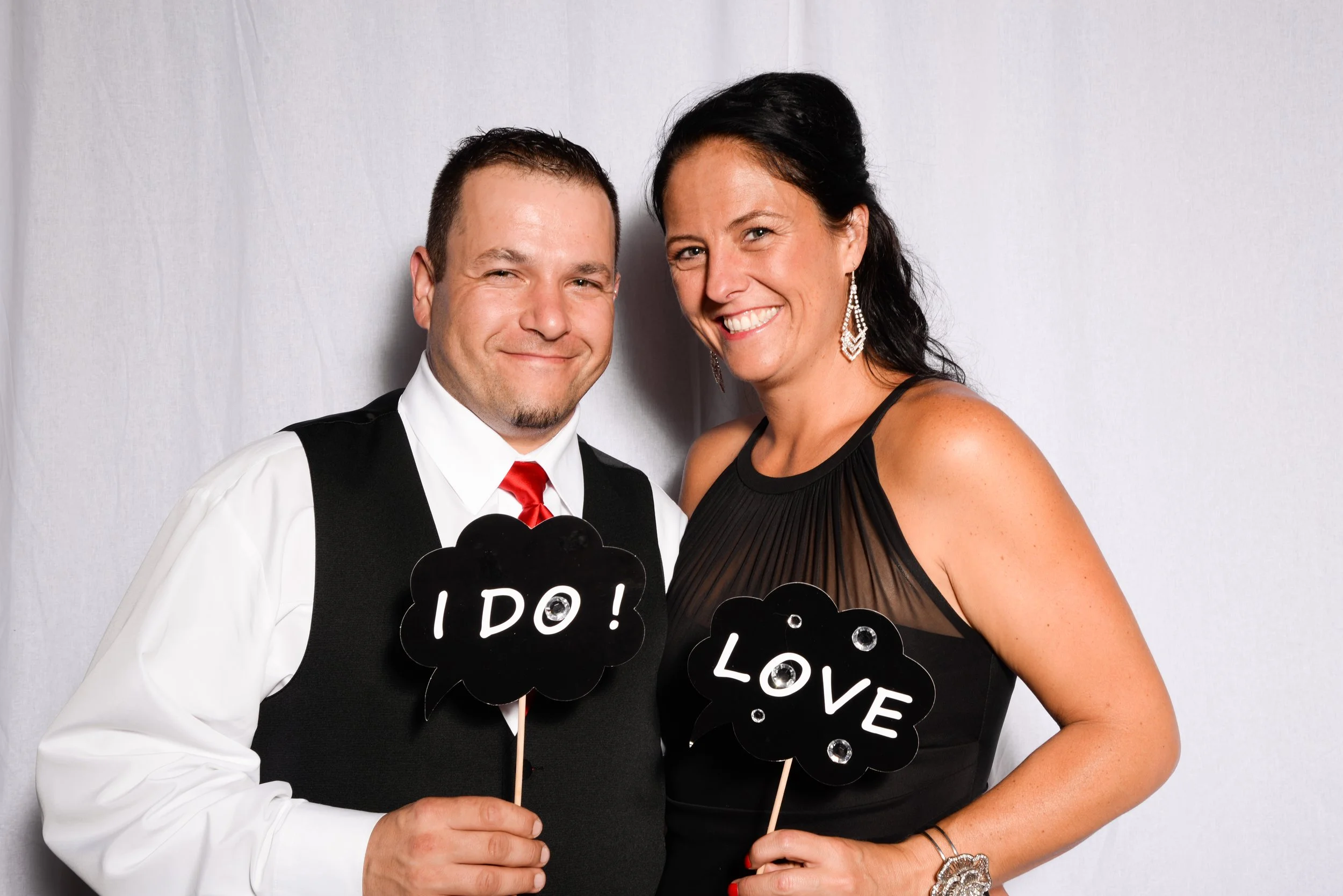 Couple posing with wedding-themed props; man holding "I Do" sign, woman holding "Love" sign, dressed in formal attire.