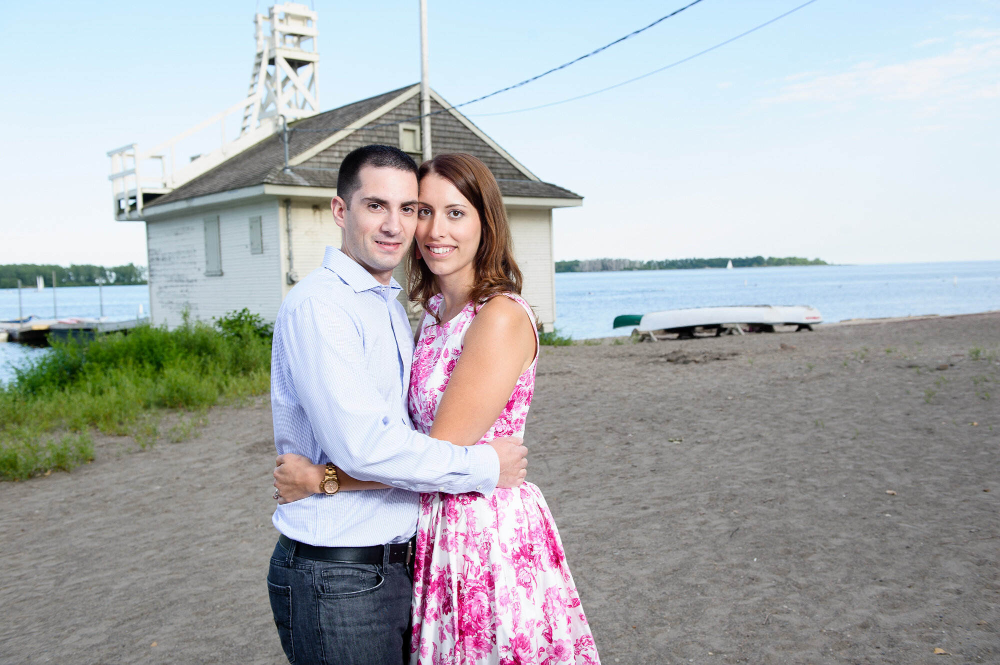 Couple embracing on a beach near a white building, with lake and boats in the background.