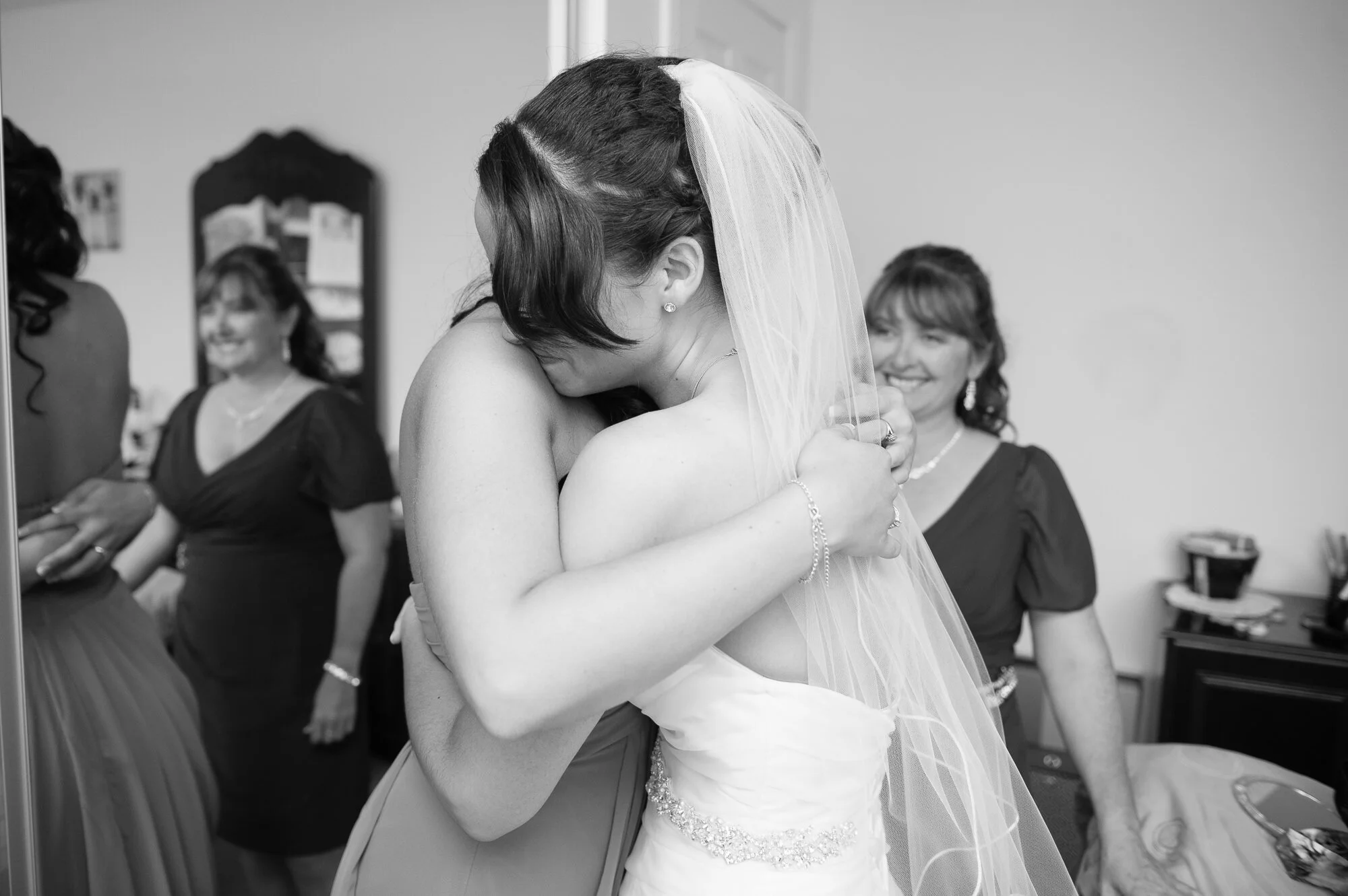 Bride in white dress hugging a woman, with smiling women in the background, in a dressing room setting.