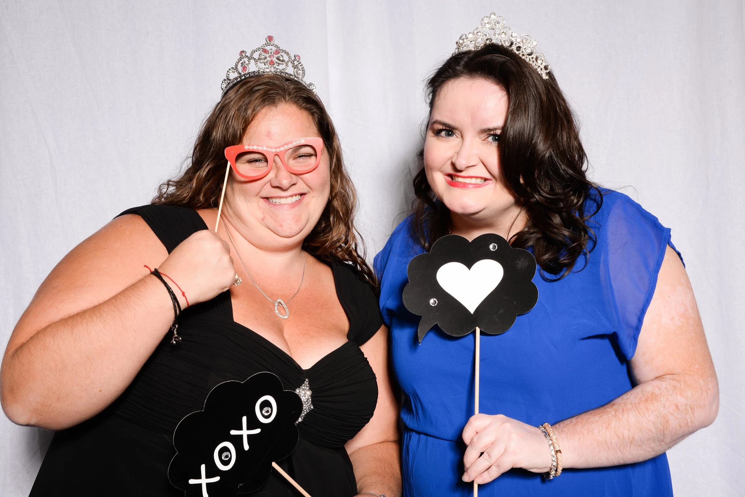 Two women with tiaras in photo booth, holding props; one in red glasses with "xoxo," the other with a heart symbol.