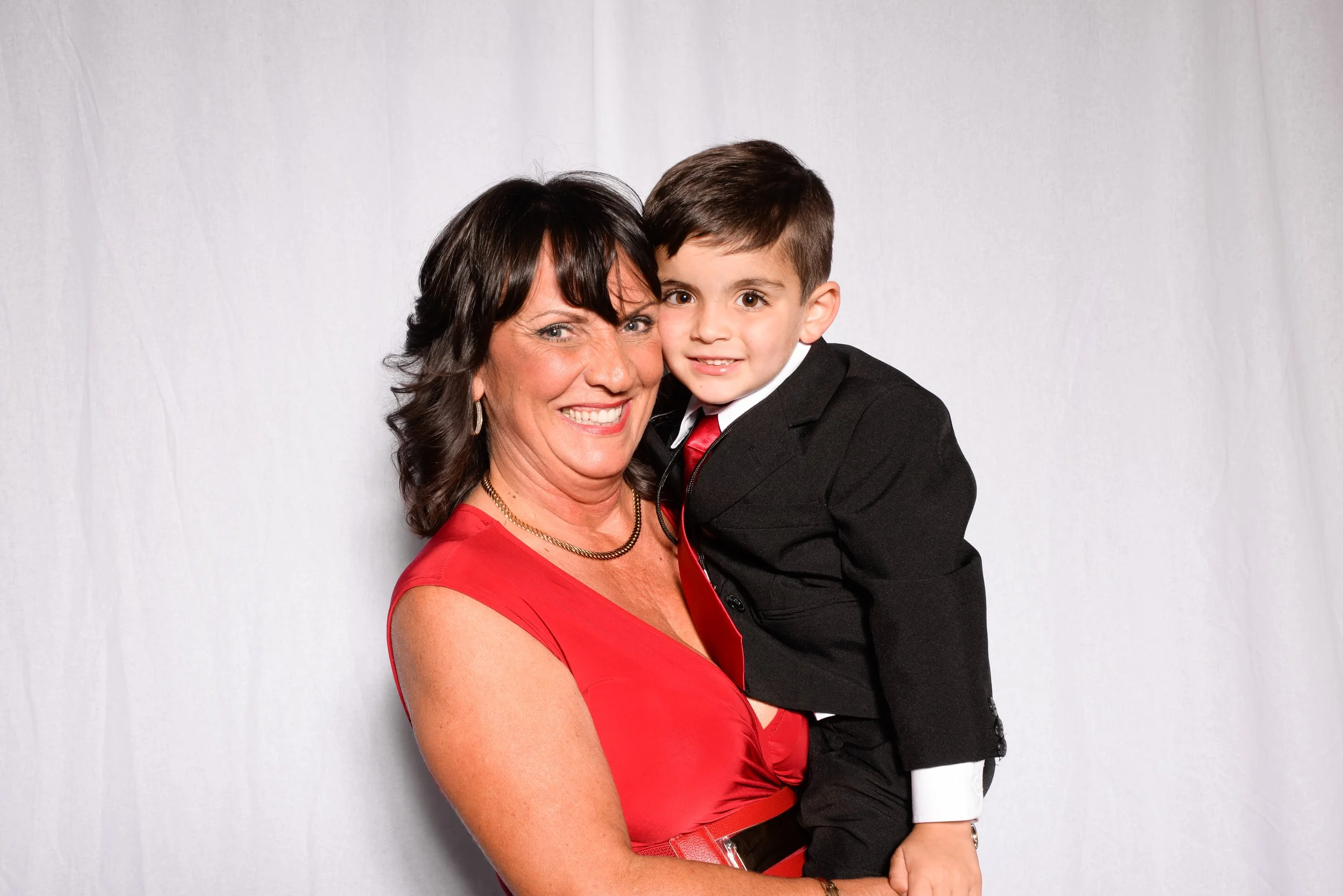 Woman in a red dress holding a young boy in a black suit and red tie against a plain backdrop.