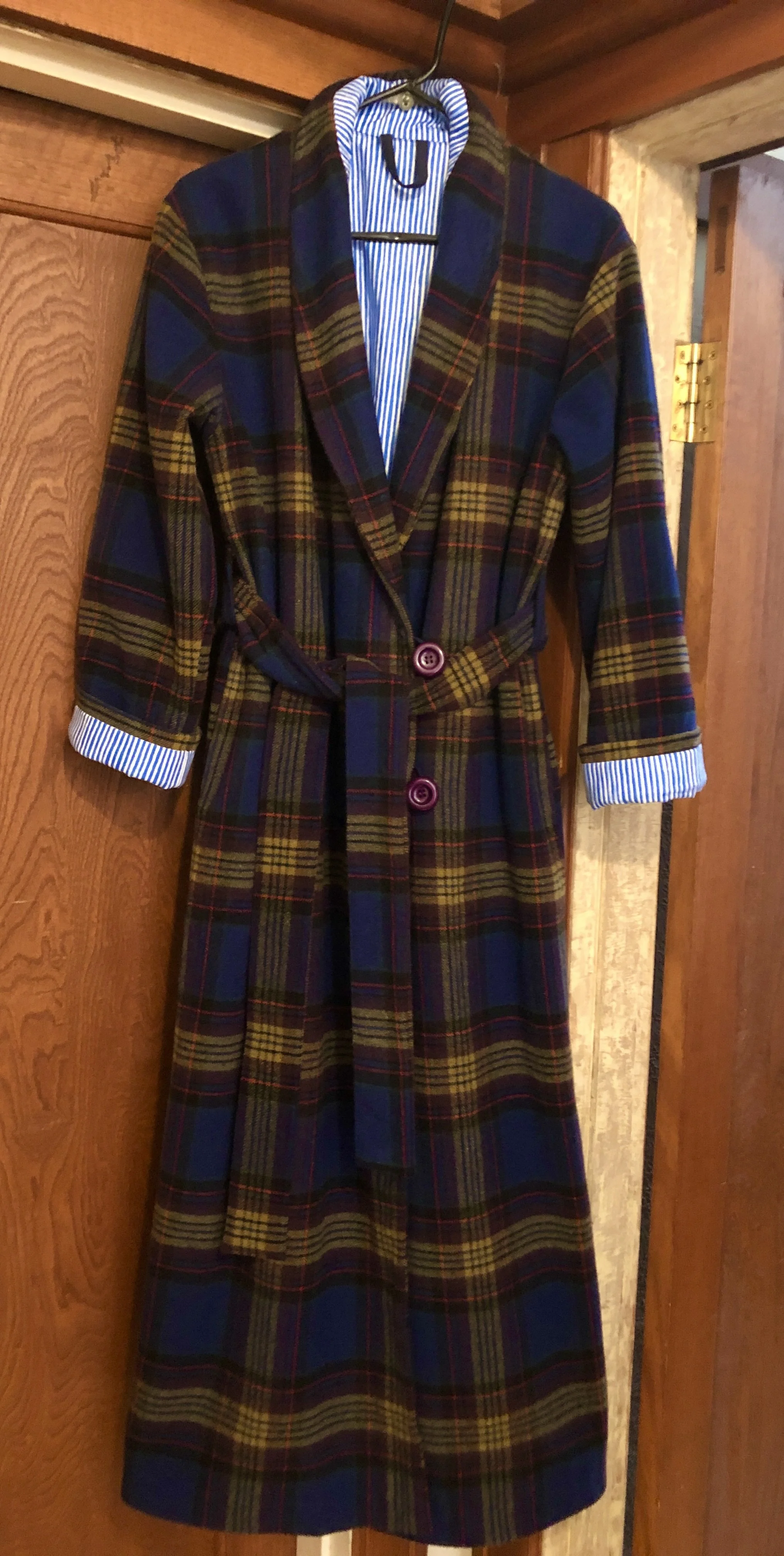 Mens wool dressing robe with a belt and large purple buttons hanging on a black hanger on a wooden wall.