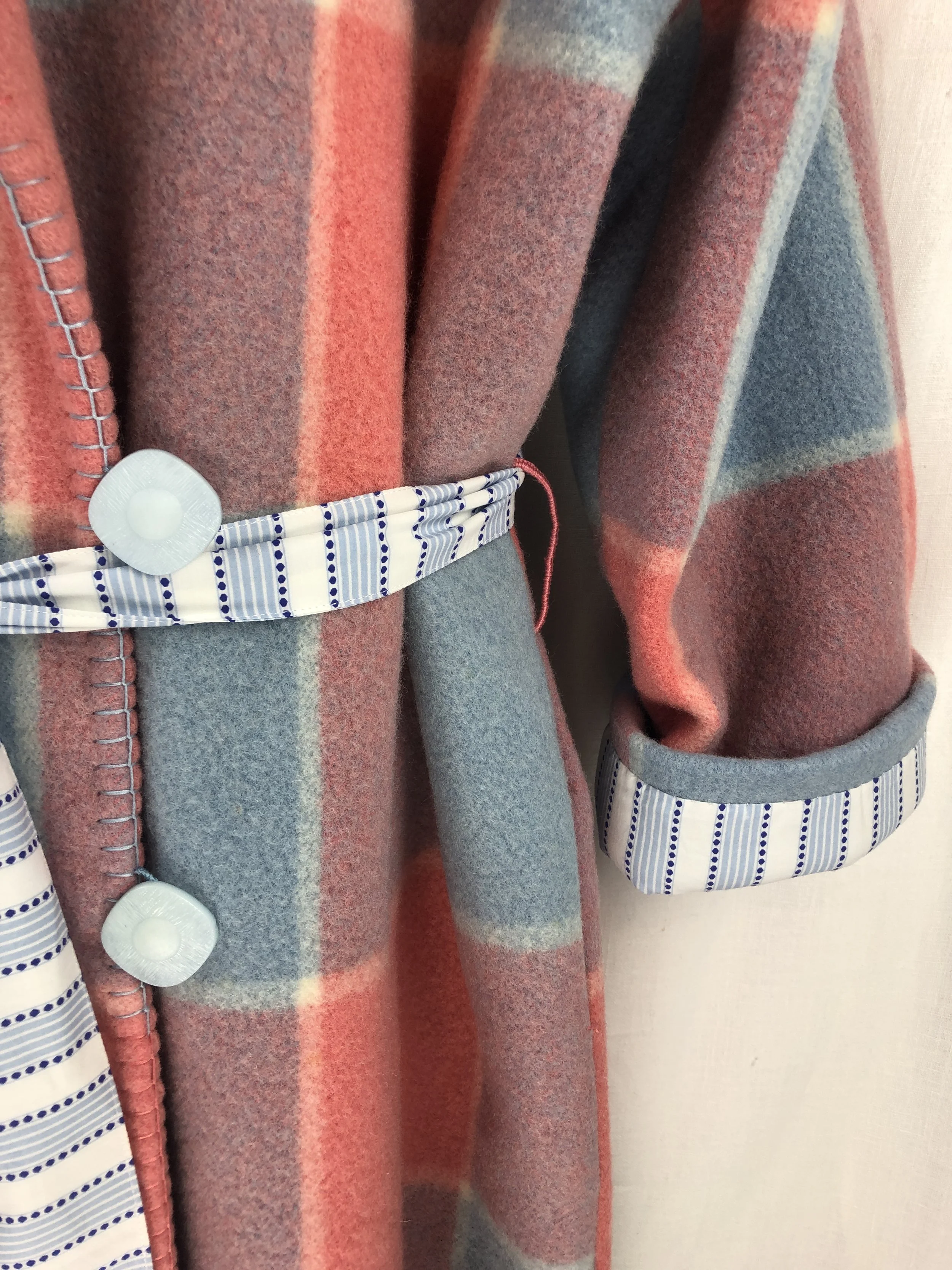 Close-up of a handmade blanket dressing gown with a mix of rust, gray, and blue colors, featuring decorative trim with striped and dotted patterns and white buttons.