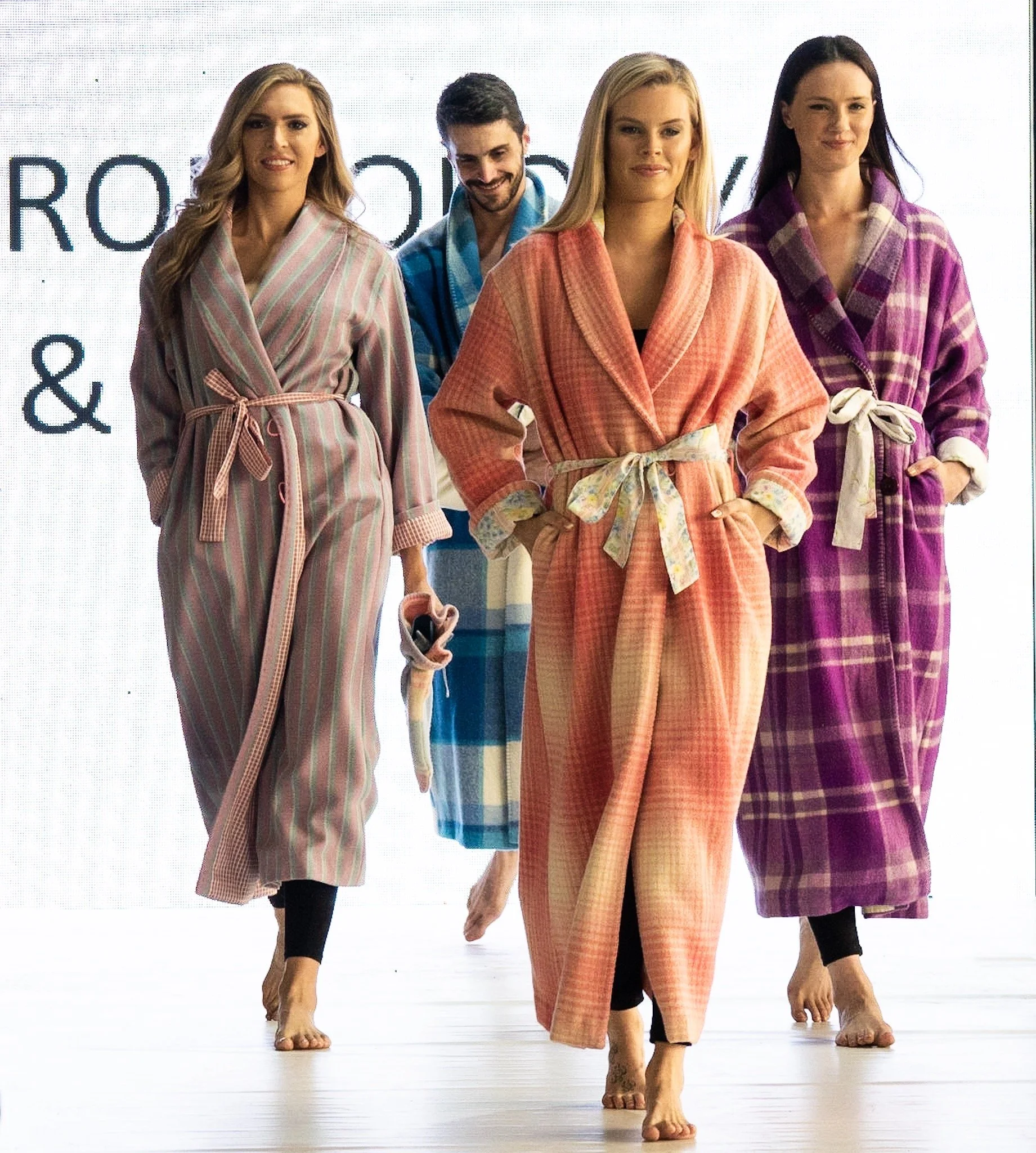 Four women and one man walking on a runway, all wearing colorful bathrobes with belts, barefoot.