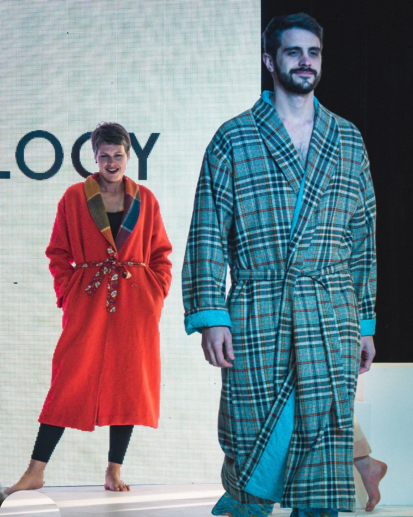 Two people on a fashion show stage wearing handmade wool bathrobes, one smiling with hands in pockets, the other with a neutral expression. From the Bendigo Woolshow, Victoria, Australia