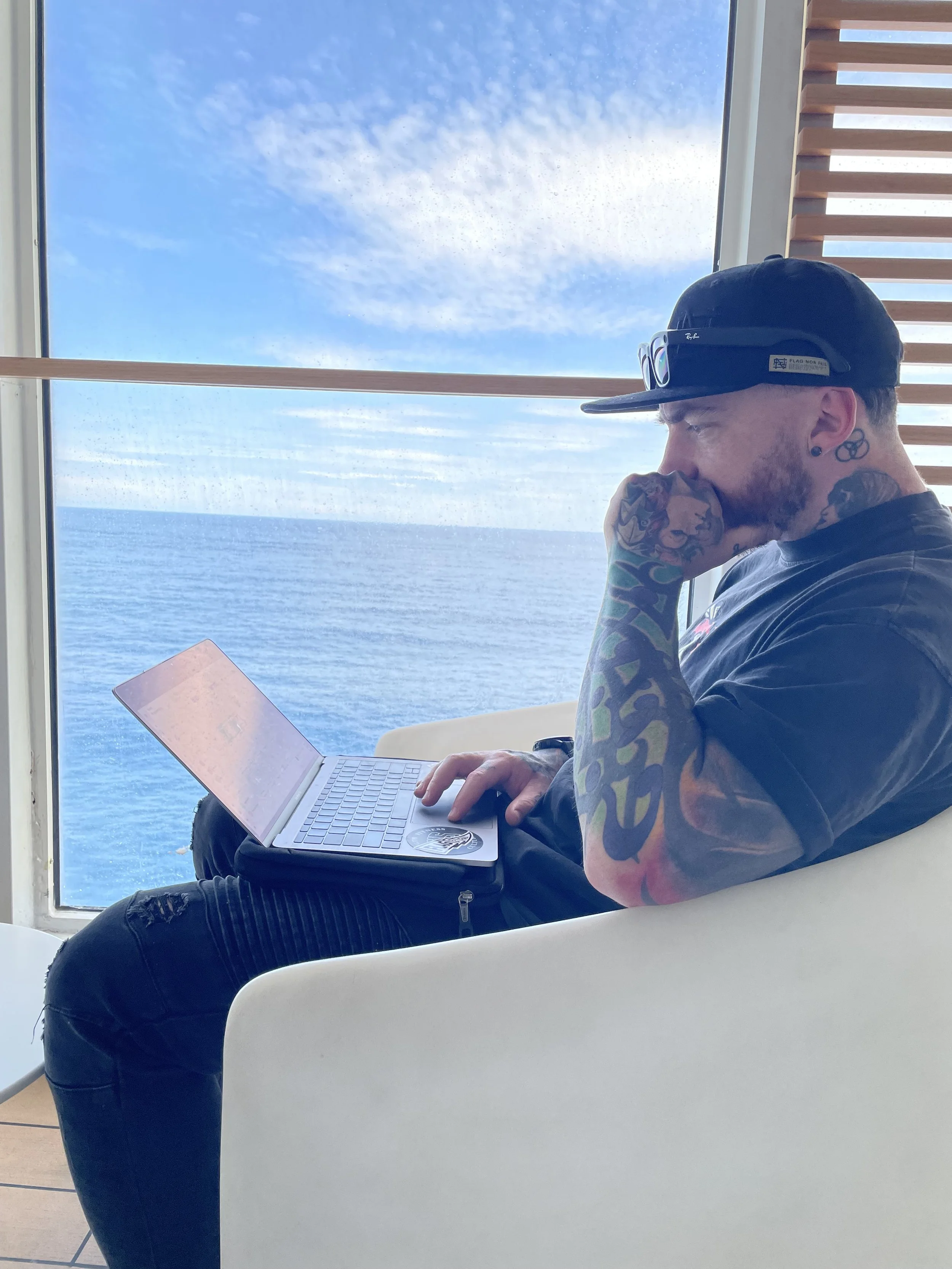 A man with tattoos and a cap sitting in a white chair, working on a laptop, with an ocean and blue sky visible through a large window behind him.