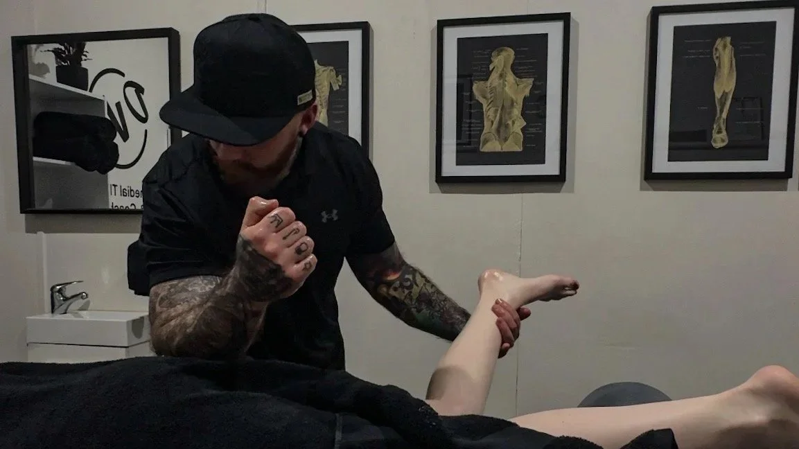 A tattooed man wearing a black cap and shirt is giving a fist bump to a person's foot while standing next to a massage table in a room with framed art on the wall.