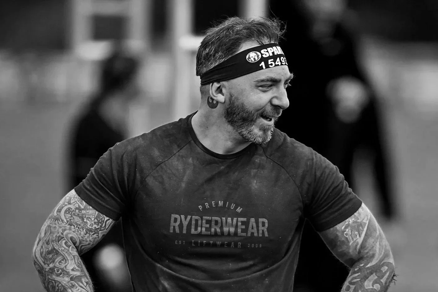 A man with a beard and tattoos on both arms wearing a black T-shirt with 'RYDERWEAR' written on it, and a black headband, smiling during a workout session.