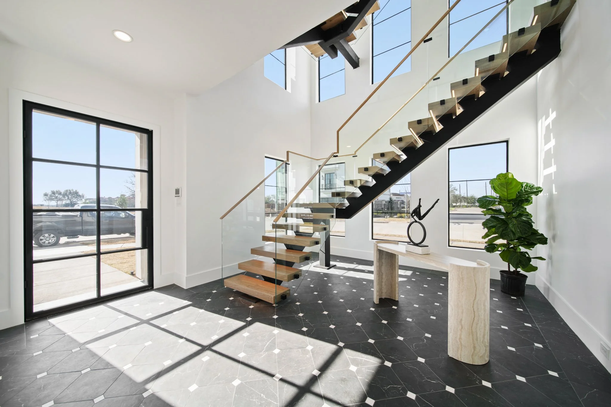 Modern interior entryway with black and wood floating staircase, large windows, potted plant, and geometric black and white tiled floor.