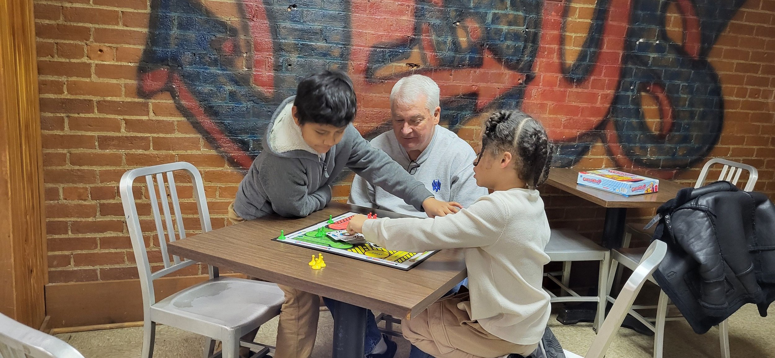 Three people playing the board game Sorry! at a table in a restaurant or cafe with a brick wall and a large painted mural of a face in the background. Two children and an older man are focused on the game, with one child reaching across the board.