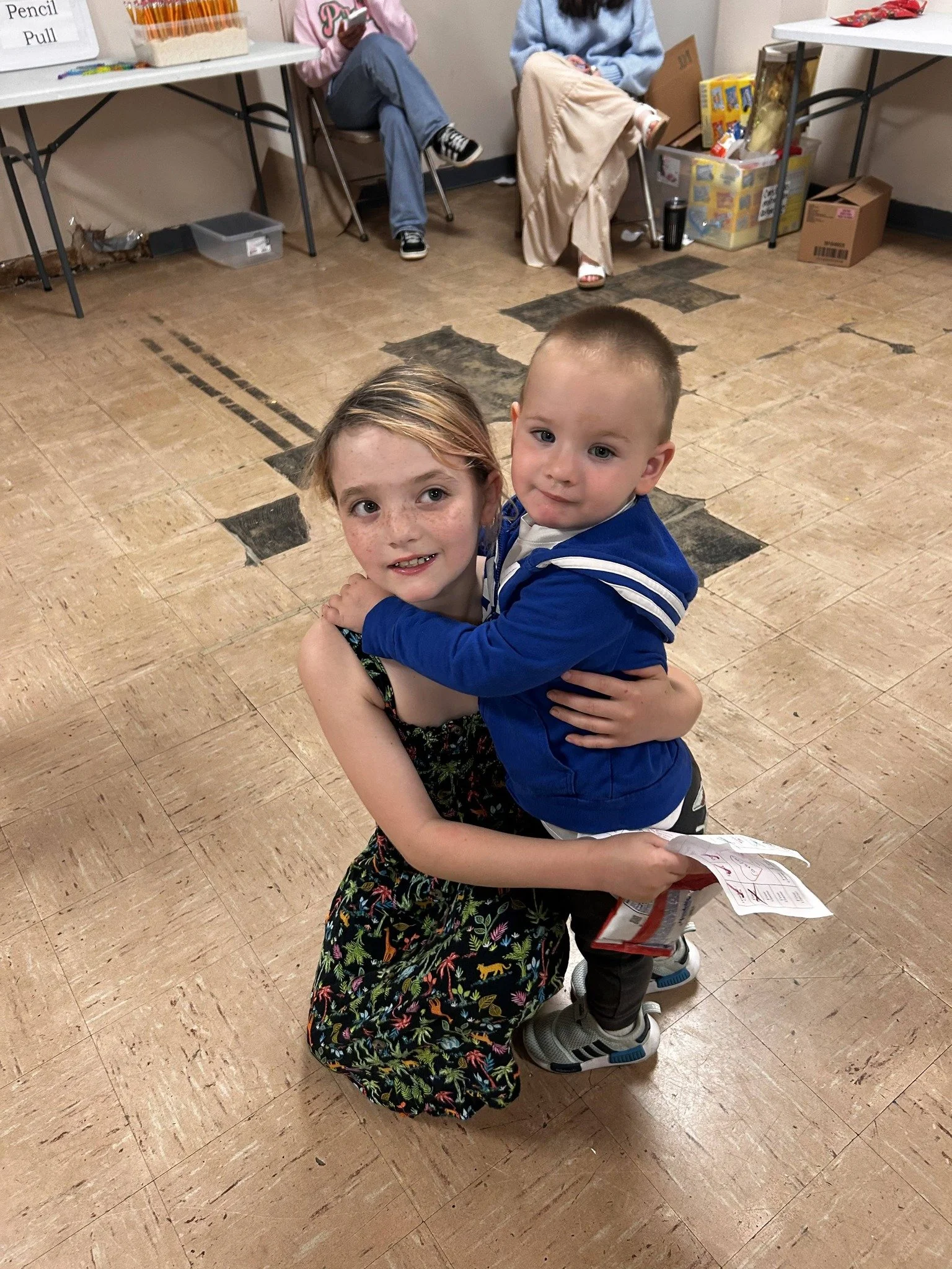 Two young children hugging each other in a room with a tiled floor and some tables and chairs in the background.