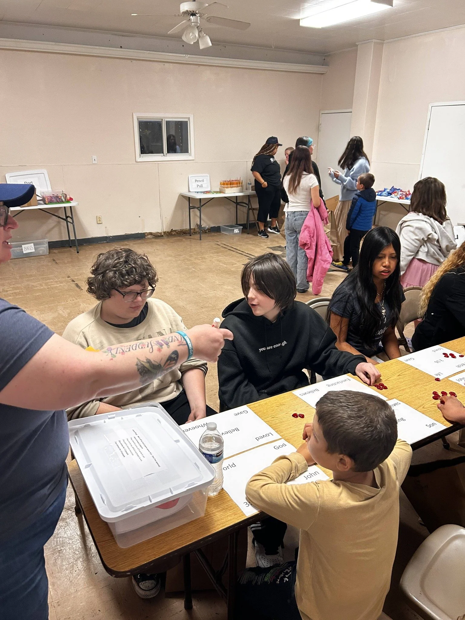 Group of children and adults seated at a table playing a game involving red chips, with word cards on the table, in a room with beige walls and a white ceiling with a light fixture. A table with snacks and signs is visible in the background.