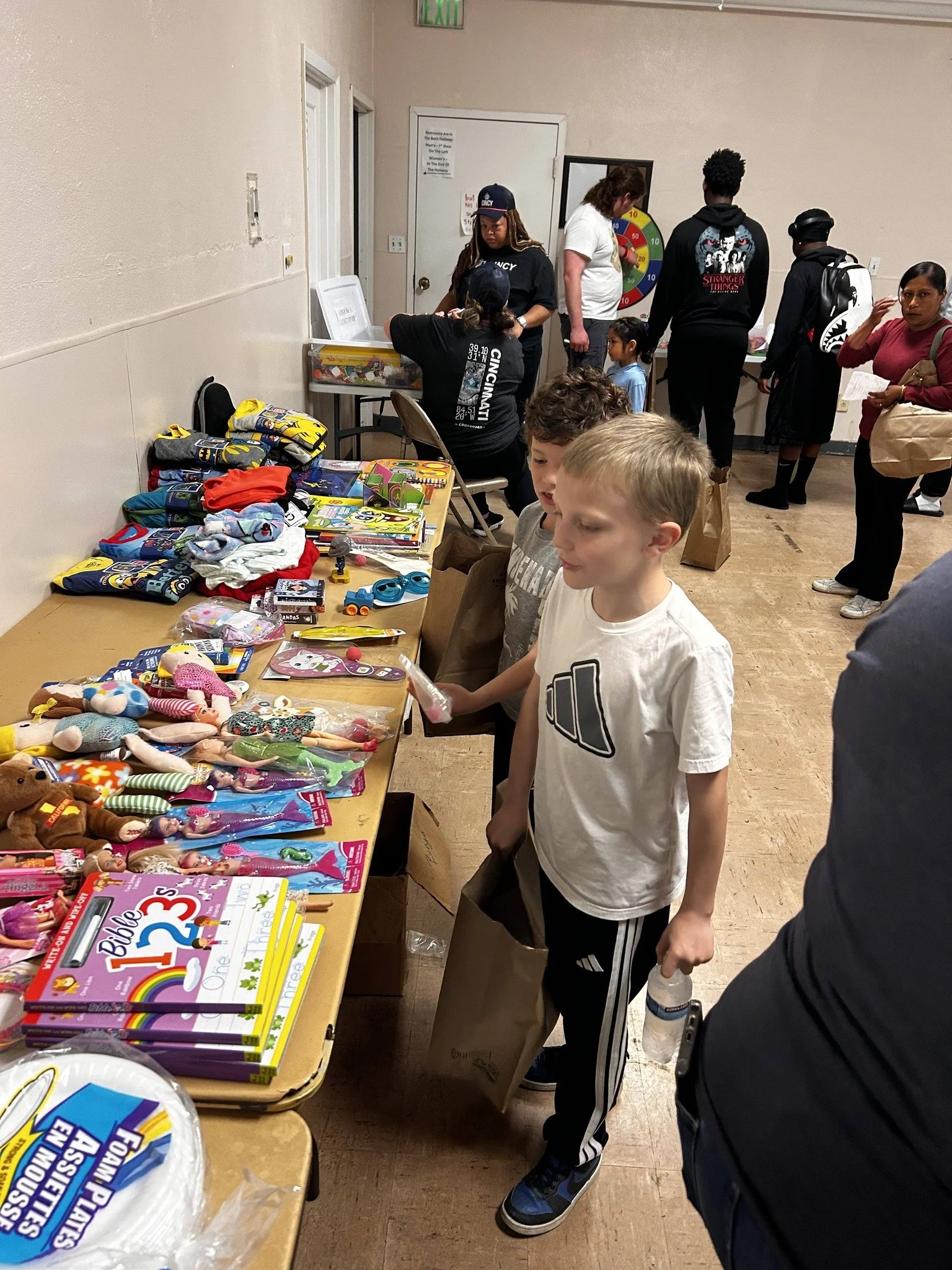 Children and adults browsing toys and books at a table during a community event or sale inside a room with beige walls and wooden flooring.