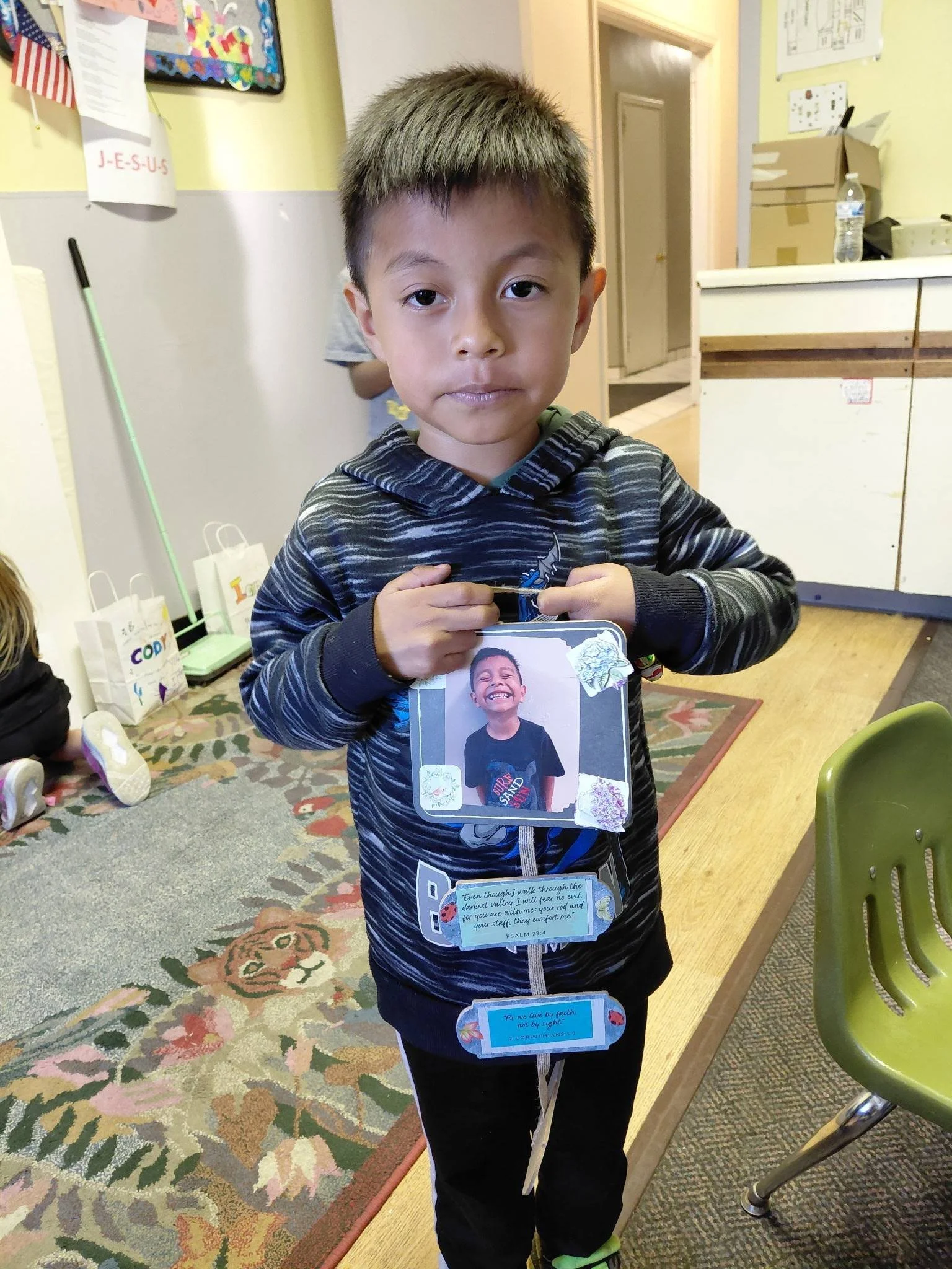 A young boy with short dark hair and a striped hoodie standing in a room, holding a laminated card with a photo of another boy smiling. Behind him is a colorful mural, a cardboard box, a water bottle, and some Christmas trees in the background.