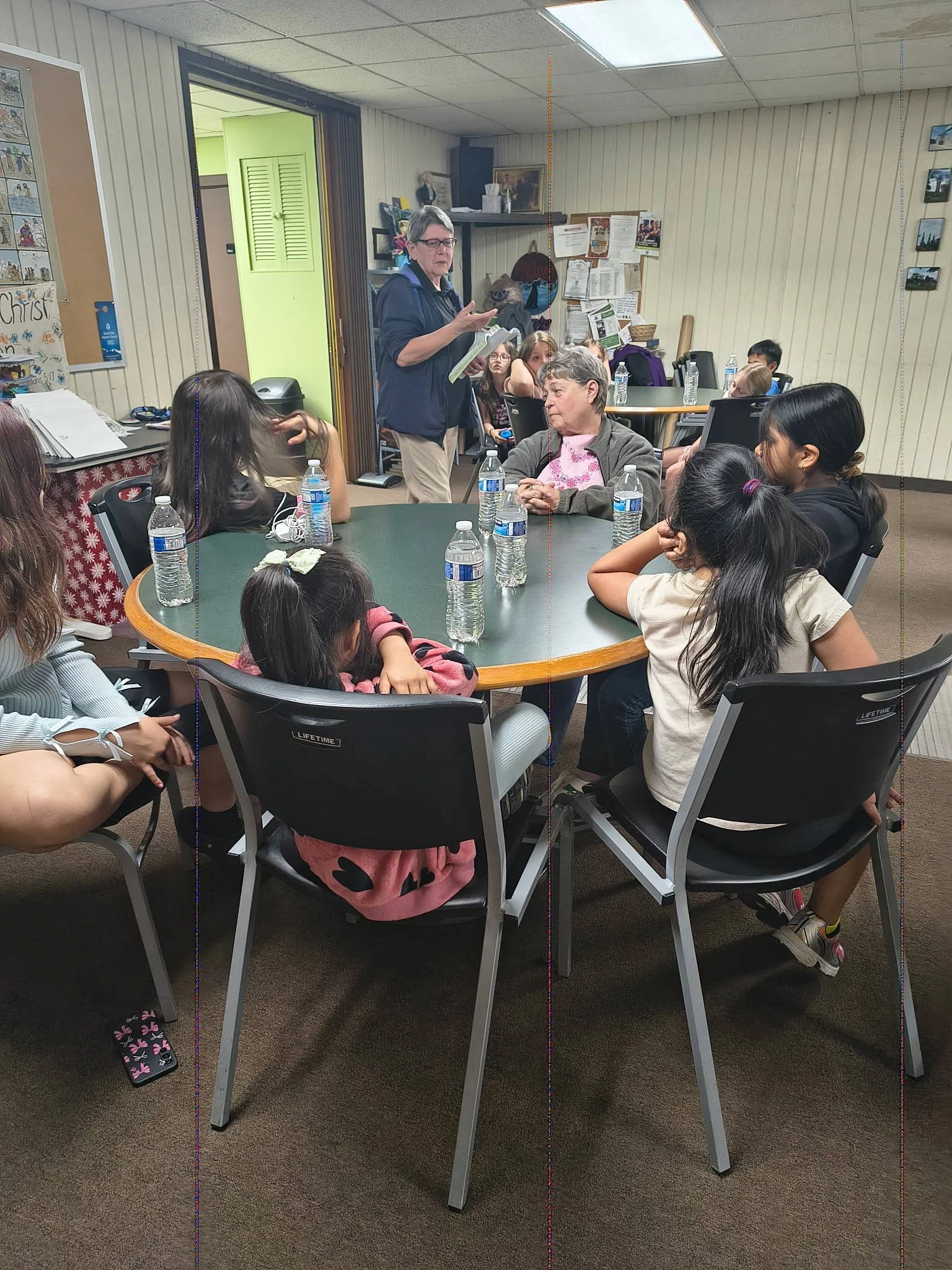A group of children sitting at a round table listening to a woman talking and holding papers, in a room with beige walls and a bulletin board.