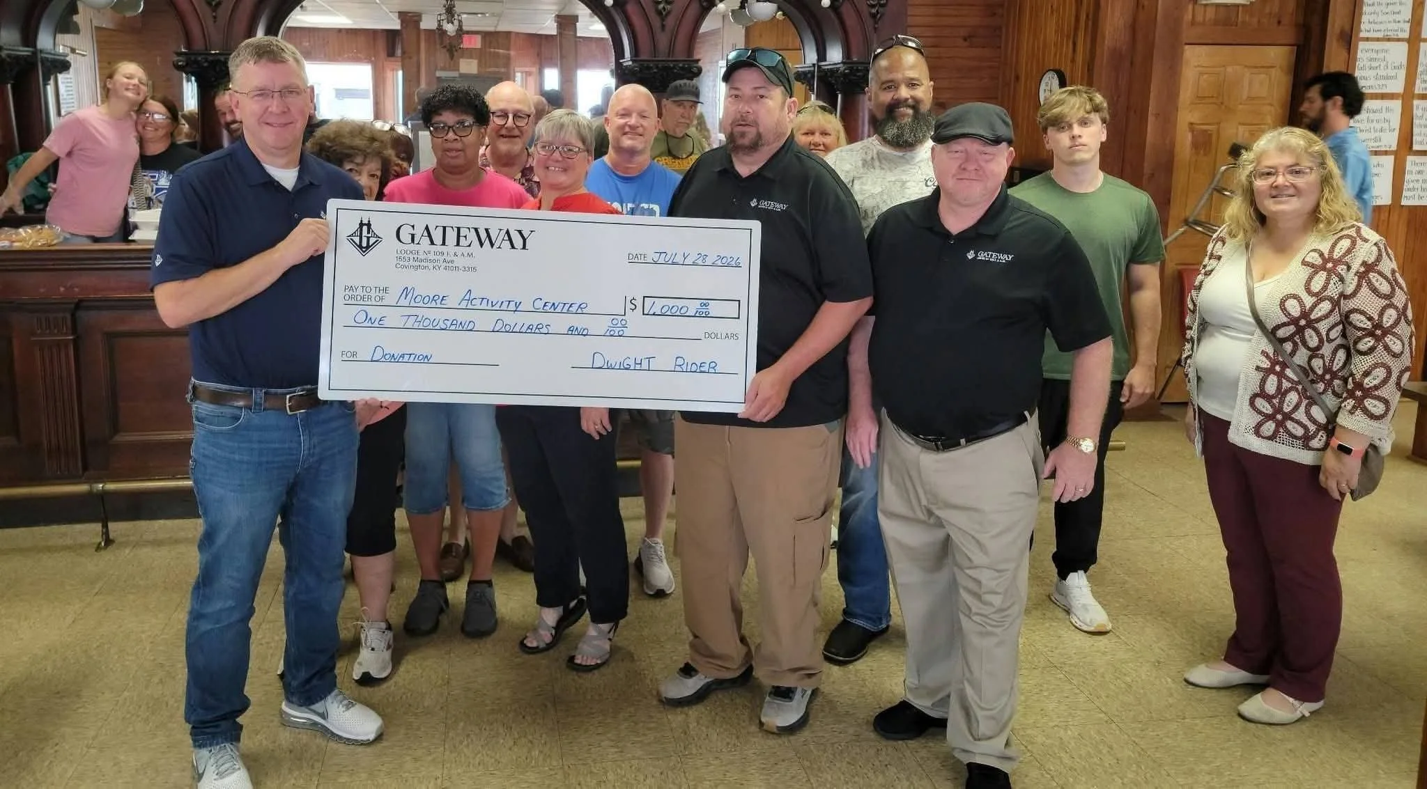 A group of people holding a large check in an indoor setting with wooden walls. The check is made out to Moore Activity Center for $1,000, signed by Dwight Rider, and dated July 28, 2026.