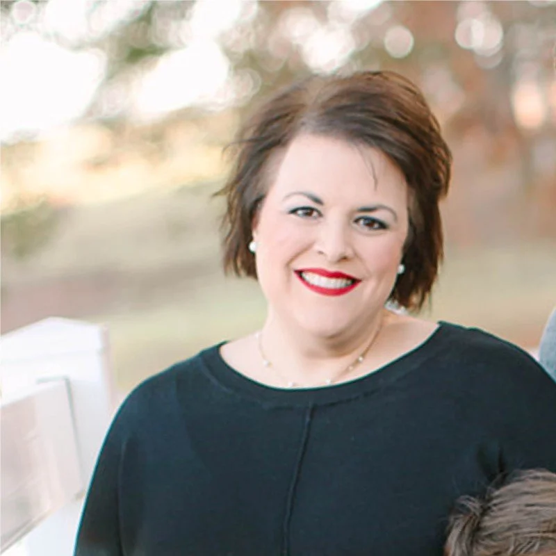 A woman with short brown hair, wearing a black top and red lipstick, smiling outdoors during daytime.