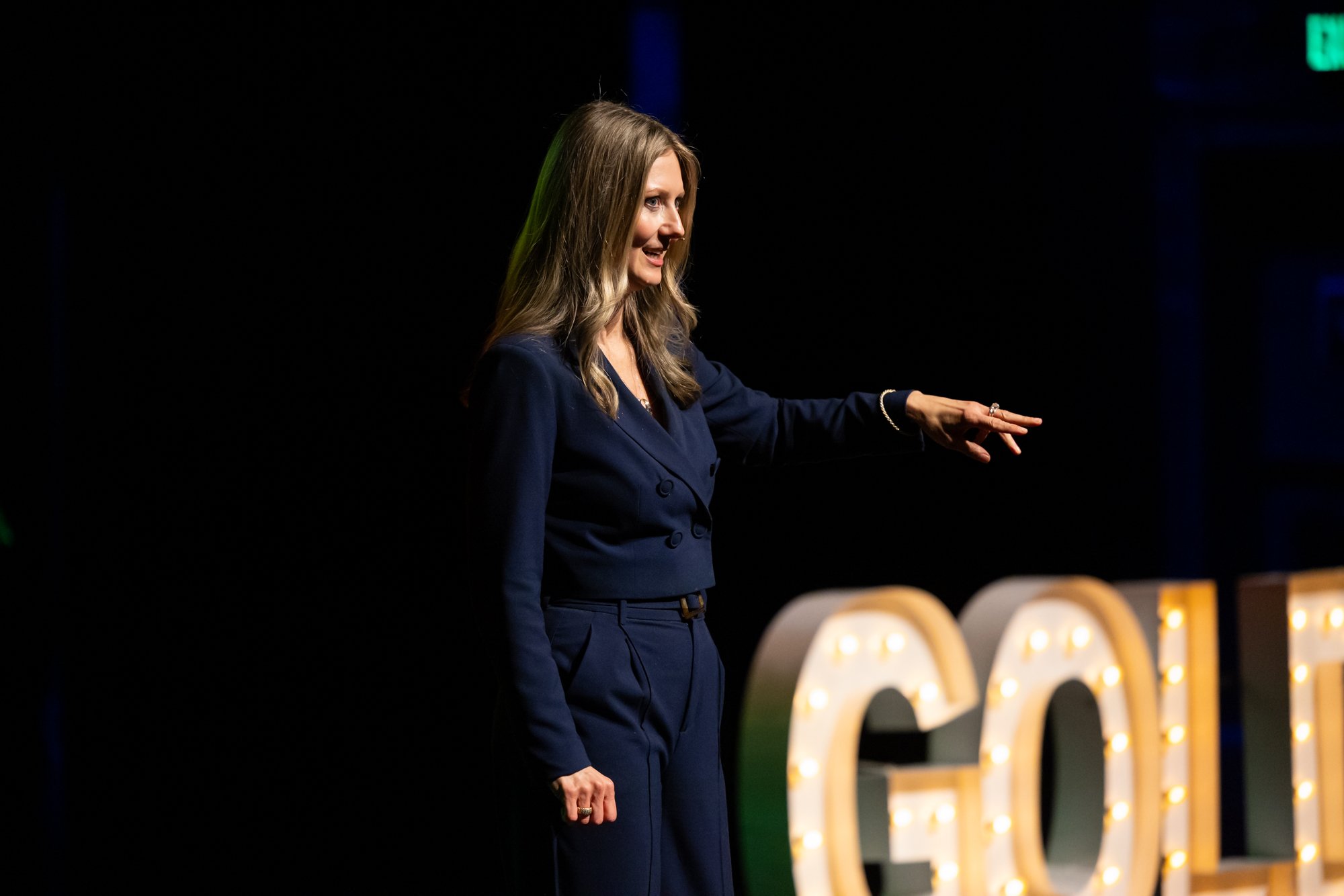Woman in a navy blue suit speaking and reaching toward an audience, while on stage with a golden, lighted sign in the background reading, "GOLD" in all caps.