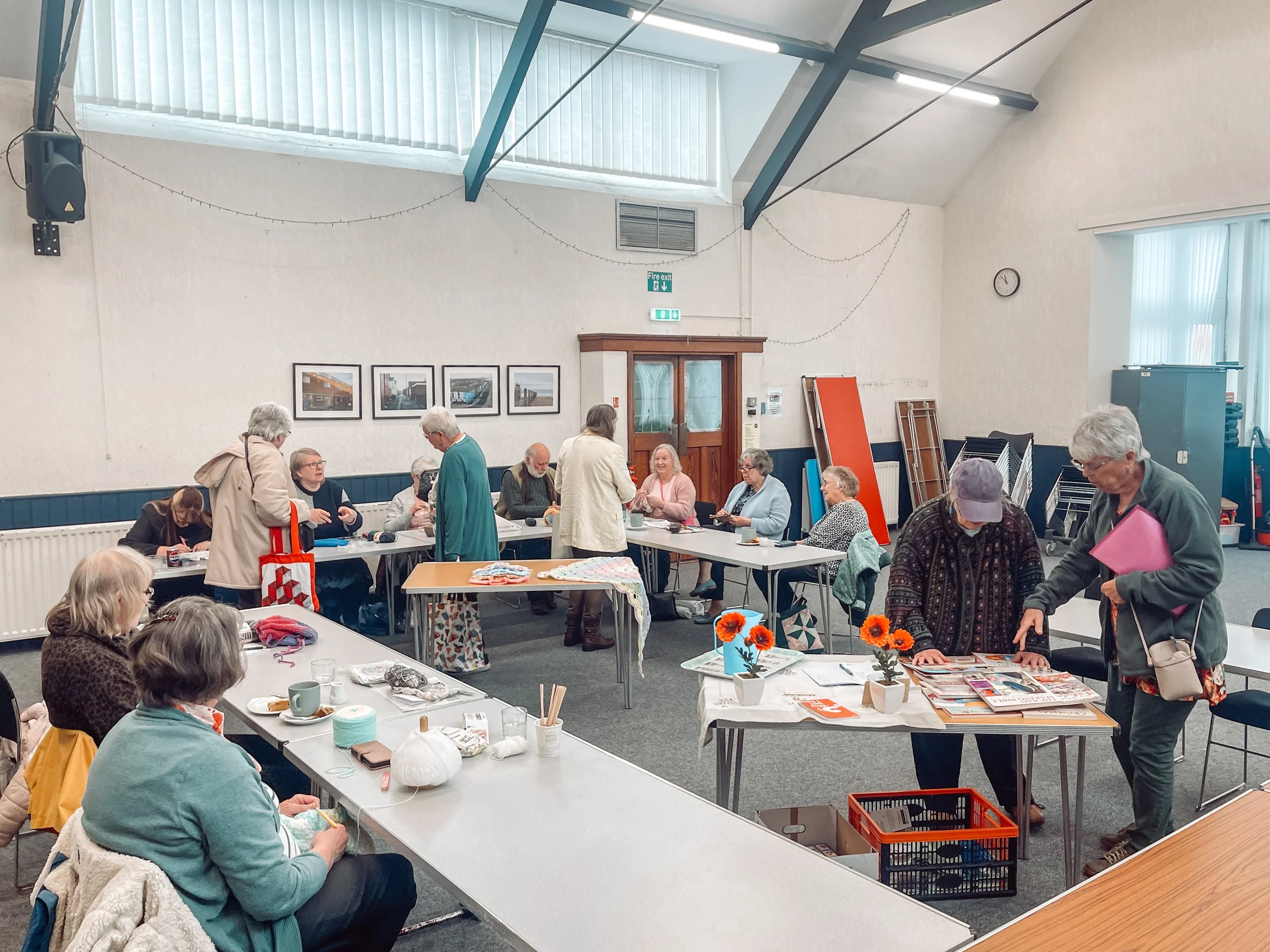 people gather around tables in a church hall to chat, eat, drink tea while working on a craft activity.