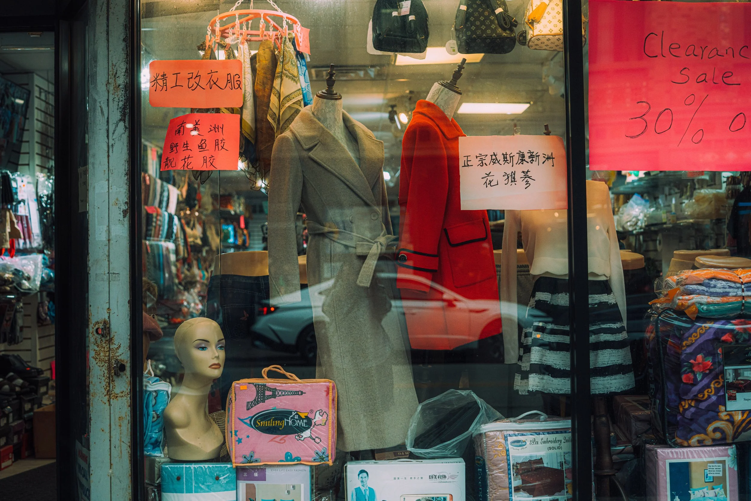 Street photograph of mannequins, clothing, handwritten signs, and merchandise filling a Chinatown, Chicago storefront window.