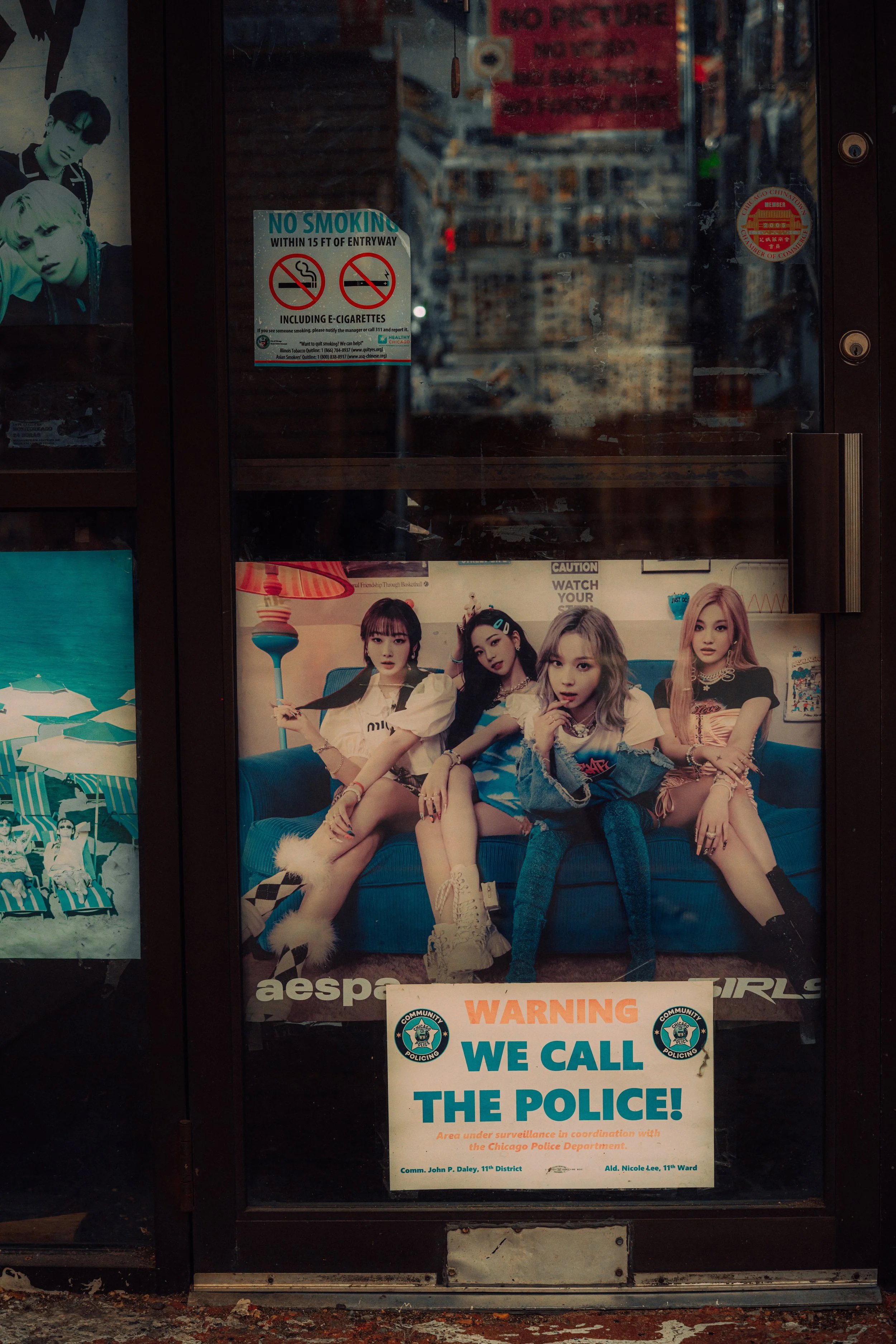 Close street photograph of K-pop posters and signs layered across a dark storefront window in Chinatown, Chicago.