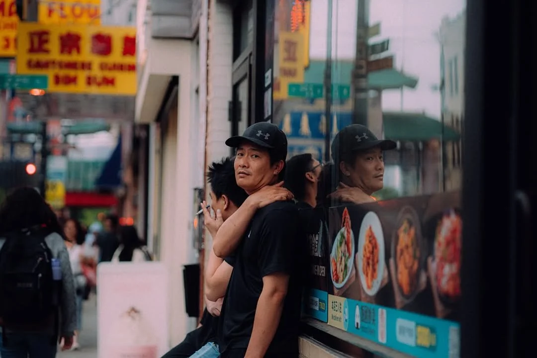 Street photograph of two men standing beside a reflective storefront window in Chinatown, Chicago, with one smoking a cigarette.
