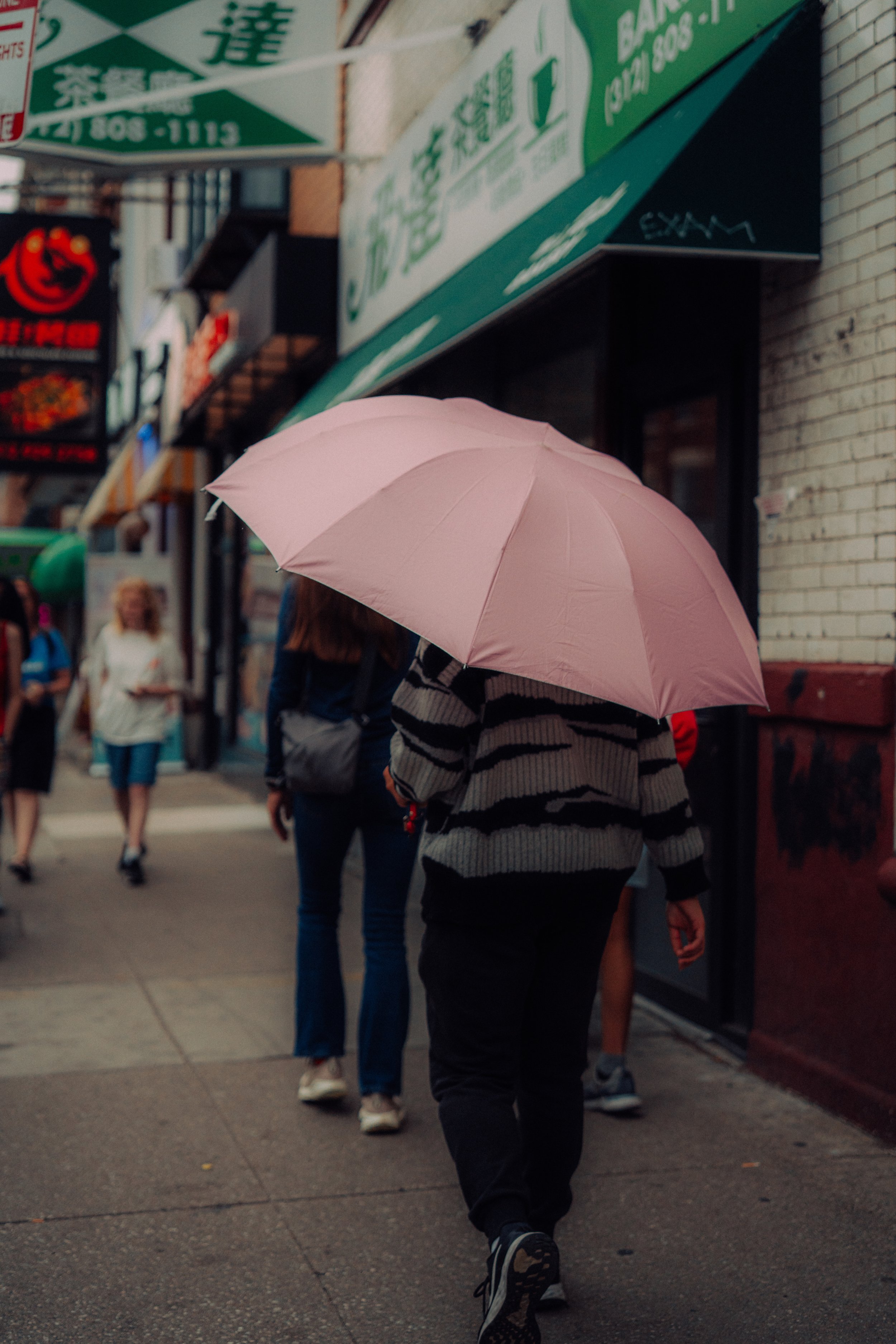 Wide street photograph of a person walking beneath a pink umbrella on a Chinatown, Chicago sidewalk.