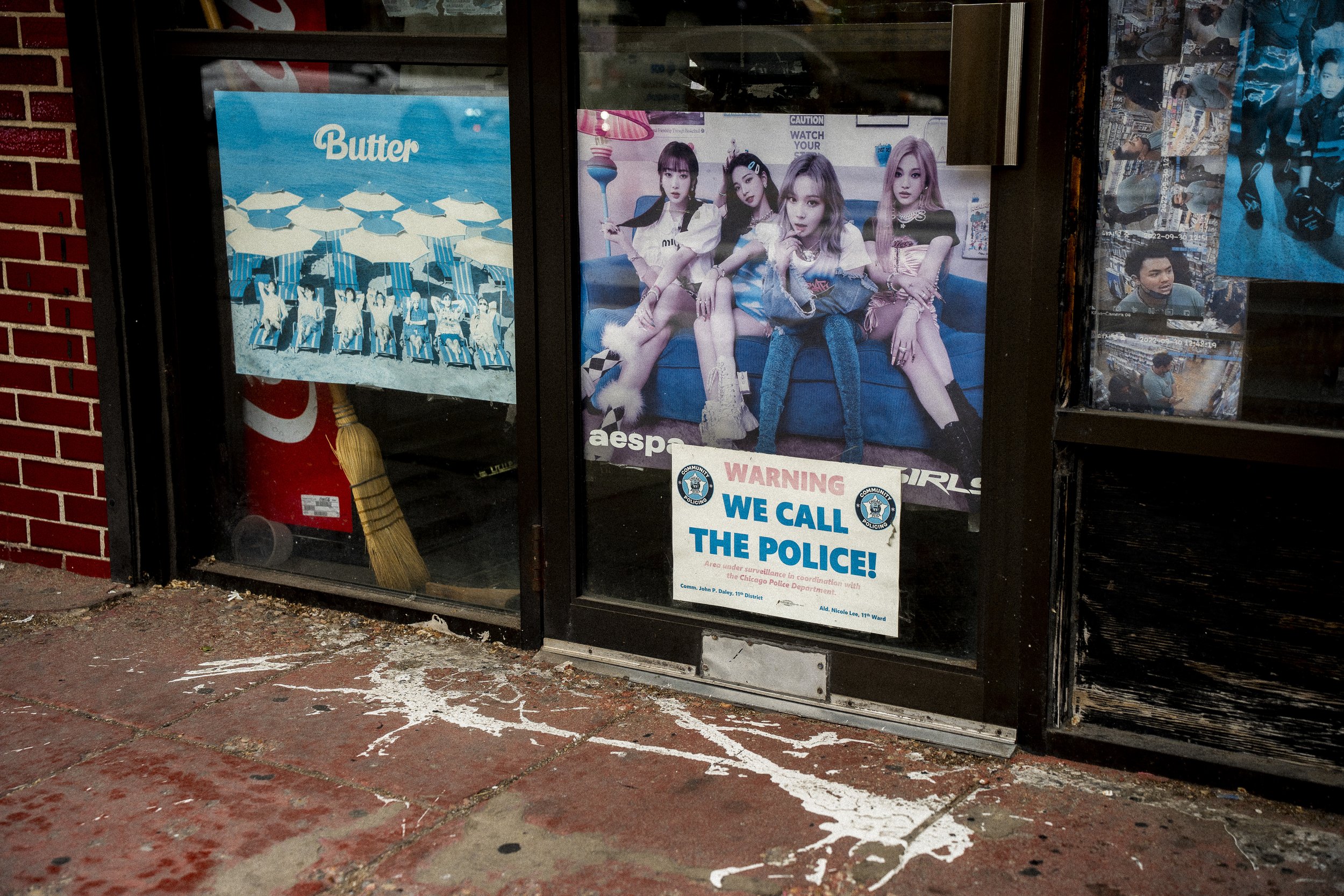 Street photograph of K-pop posters and warning signs covering a storefront window in Chinatown, Chicago.