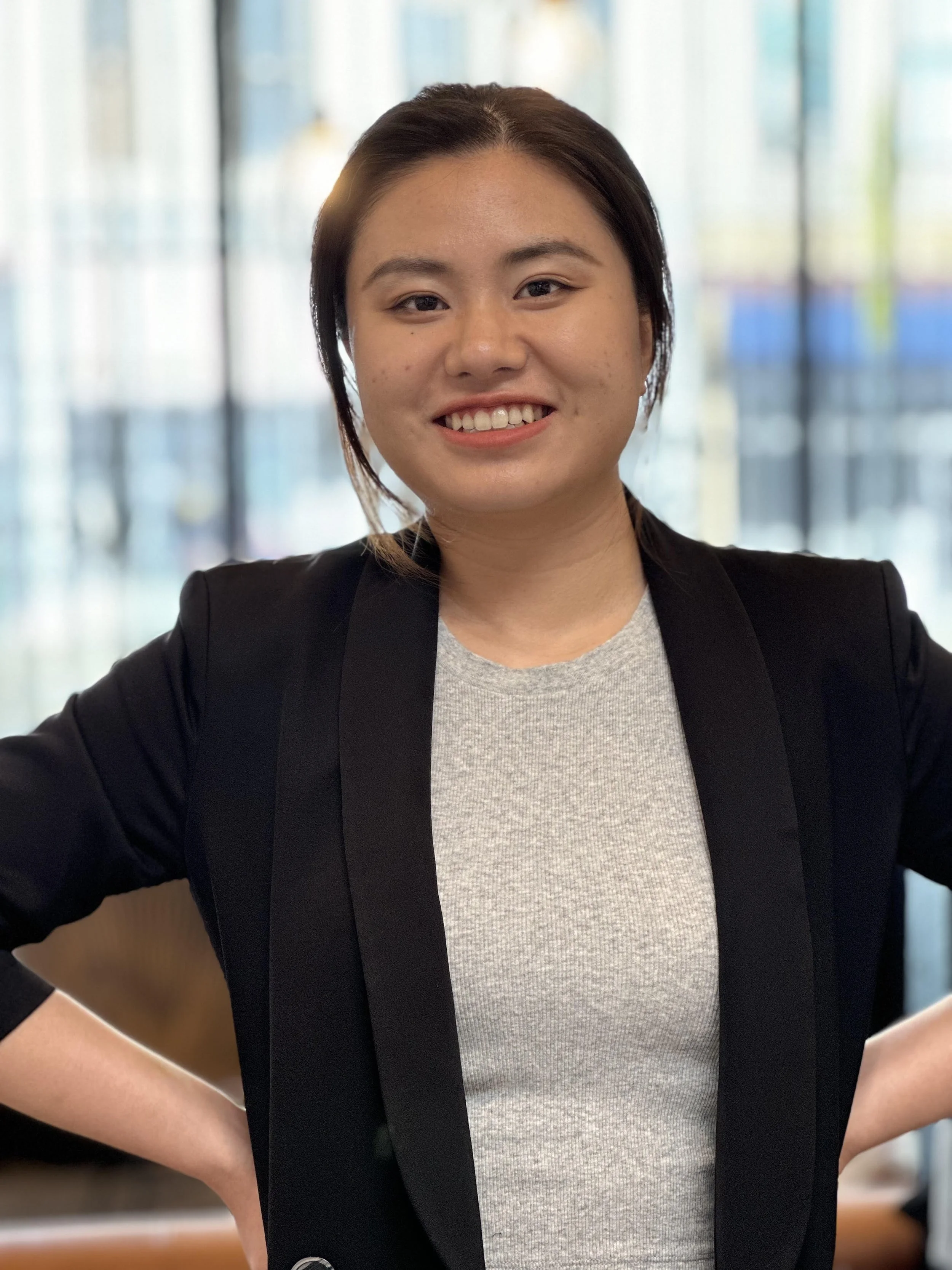 A smiling woman in a black blazer and gray shirt standing indoors with her hands on her hips, in front of a window with colorful blurred background.