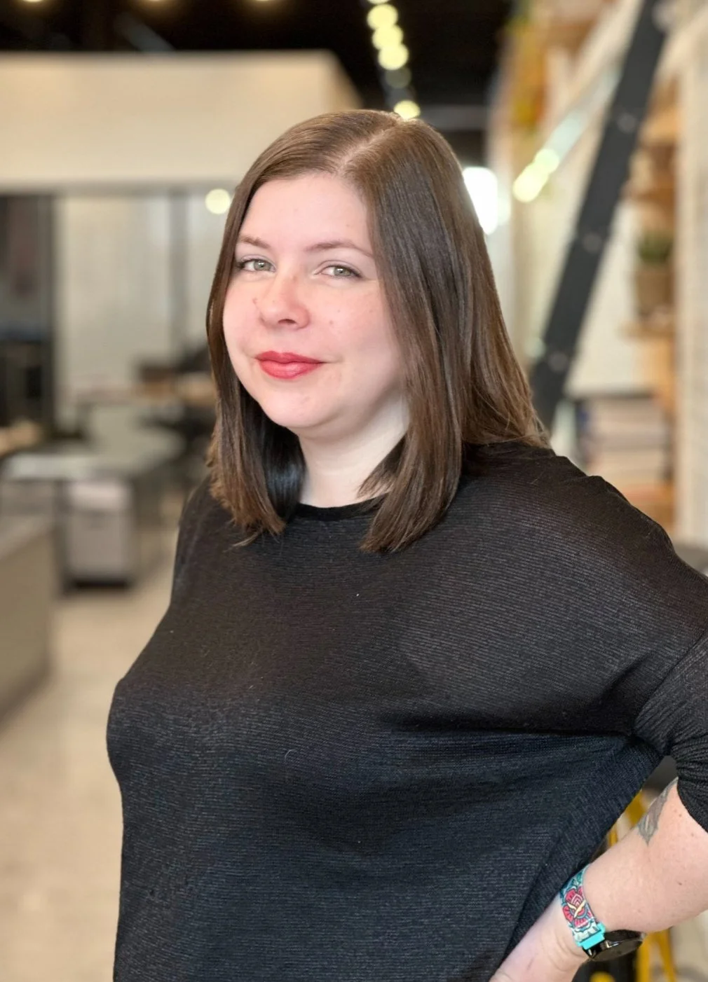 A woman with shoulder-length brown hair, fair skin, and wearing a black top, standing in an indoor space with blurred background that includes a ladder and plants.