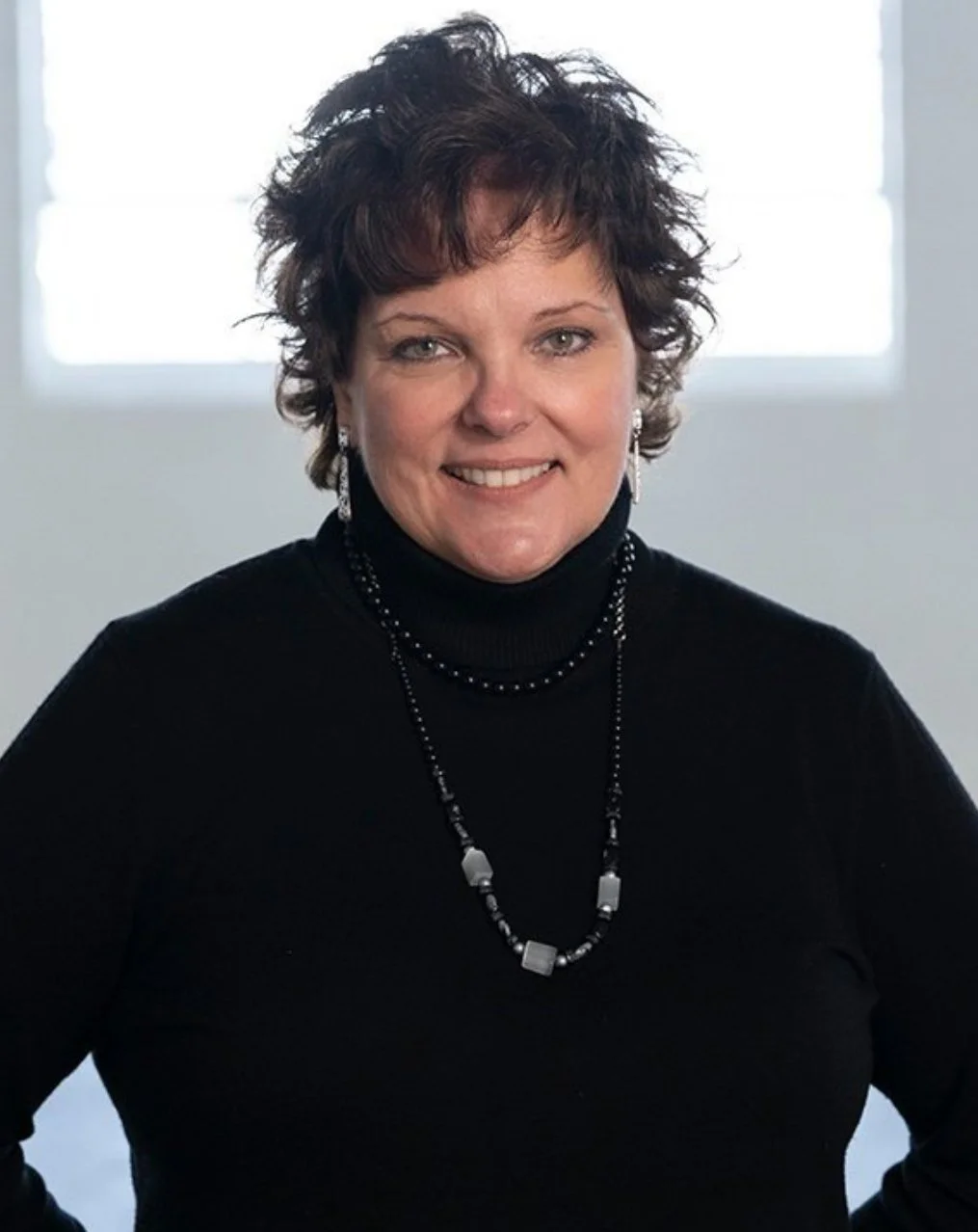 A woman with short, curly dark hair wearing a black turtleneck and jewelry, smiling in front of a bright window.