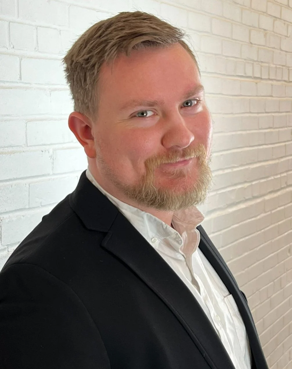 A man with blond hair, blue eyes, and a beard, wearing a black suit jacket and a white shirt, standing against a white brick wall.
